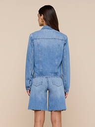 A woman with long brown hair stands facing away, wearing a slim fit Celine Jean Jacket by L’AGENCE in a light denim wash and matching knee-length denim shorts with frayed hems against a plain light background.