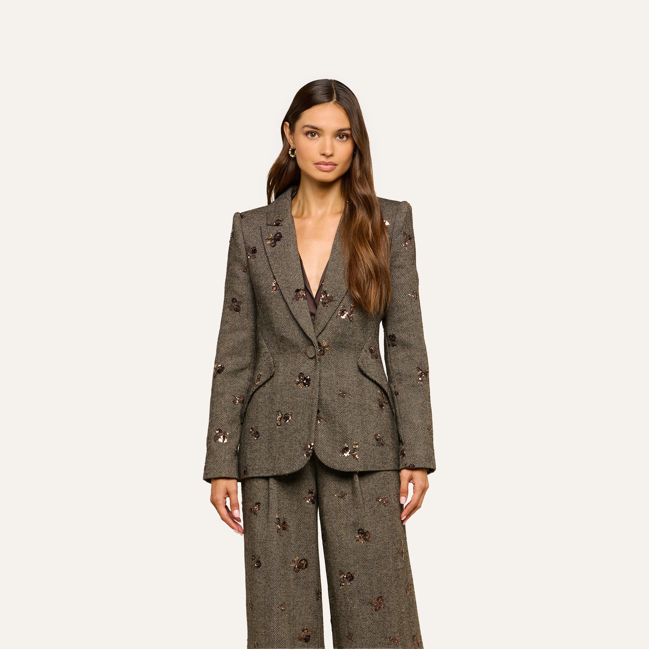 A woman wearing the Chamberlain Sequined Blazer—a gray, single-breasted piece with subtle floral embellishments—styled with wide-leg pants, stands against a plain light background.