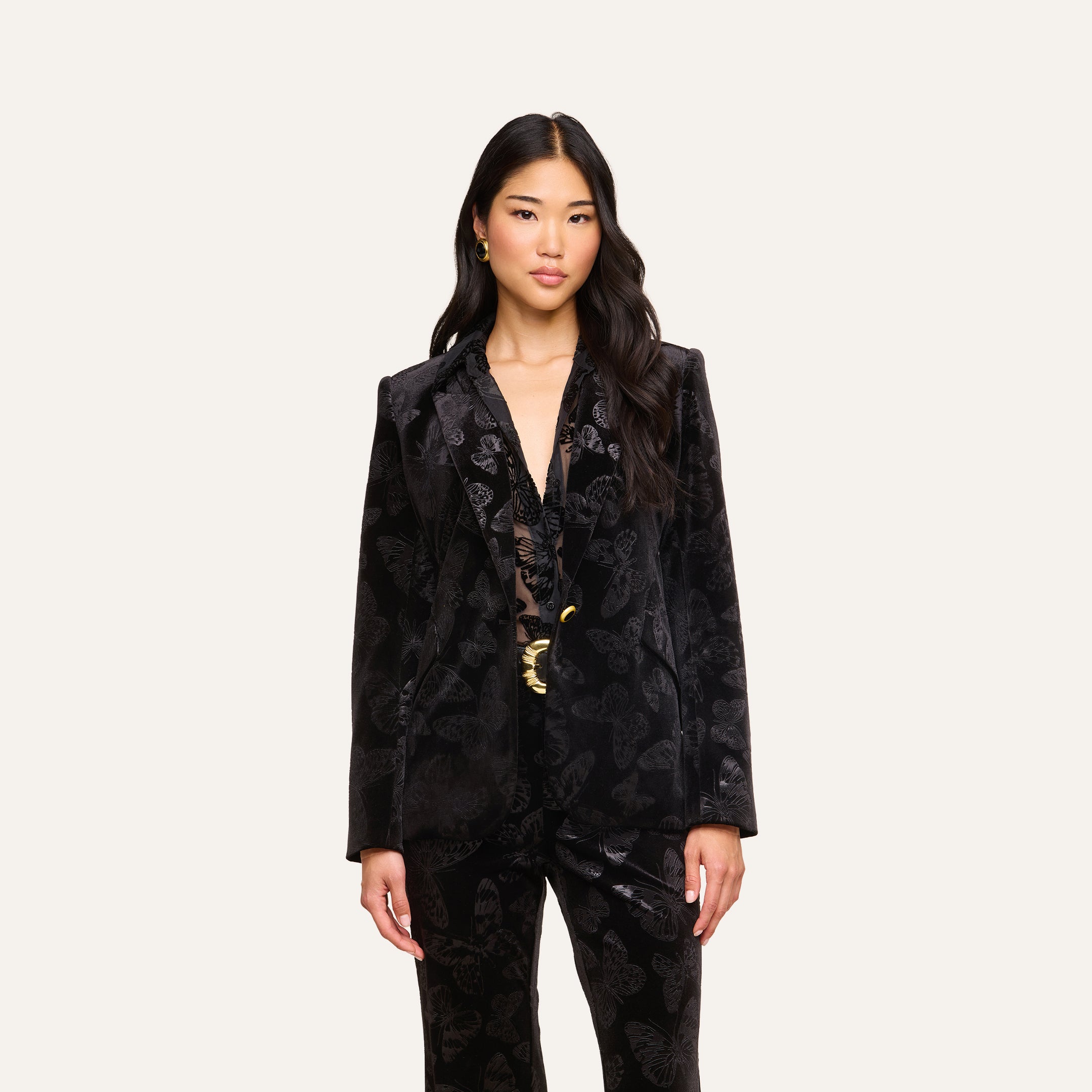 A woman with long dark hair wears the Chamberlain Velvet Blazer by L’AGENCE over a lacy top, standing against a plain light background.