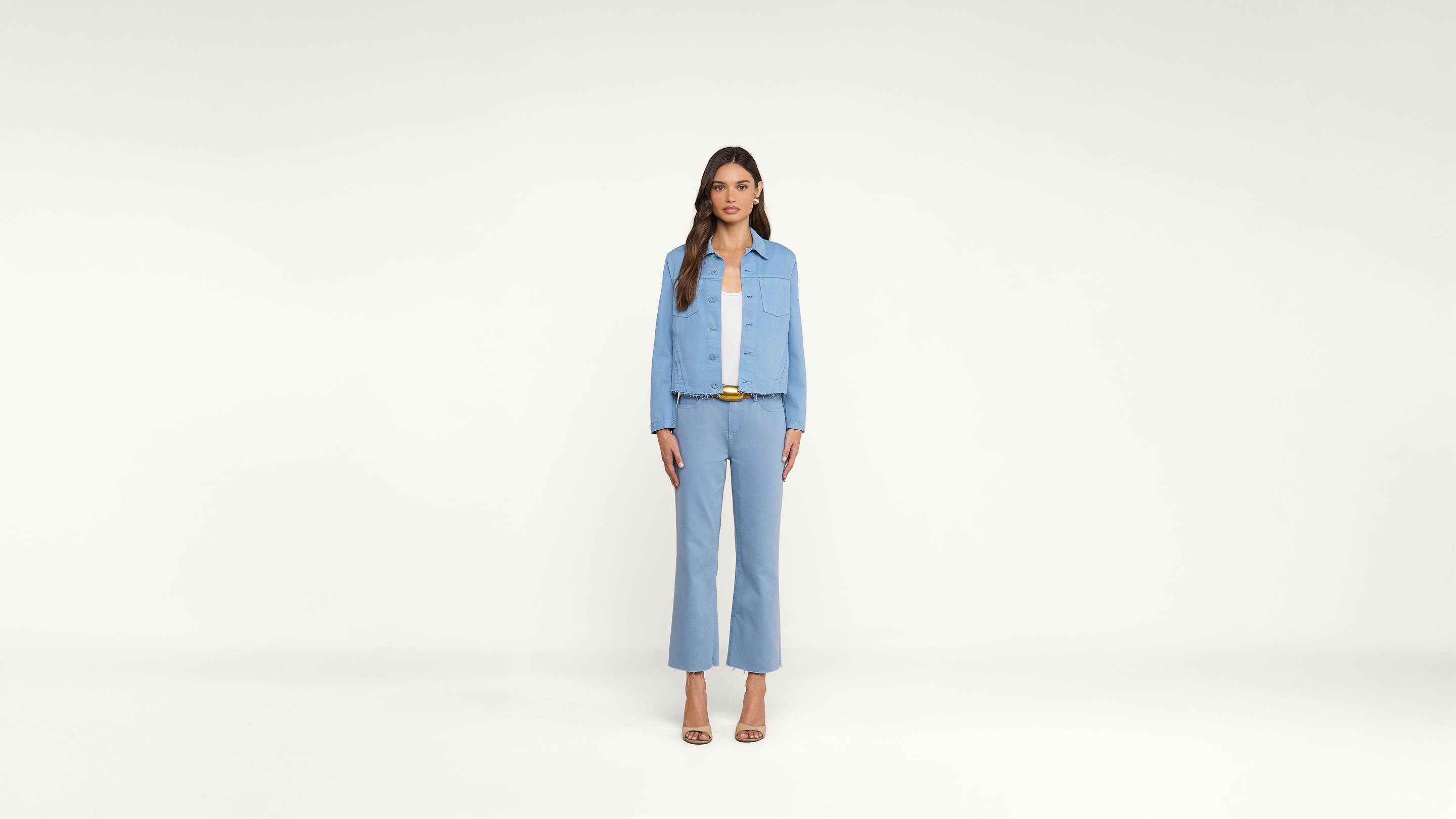 A woman stands facing forward against a plain white background, wearing a light blue jacket, matching pants, a white top, a brown belt, and beige open-toe heels.