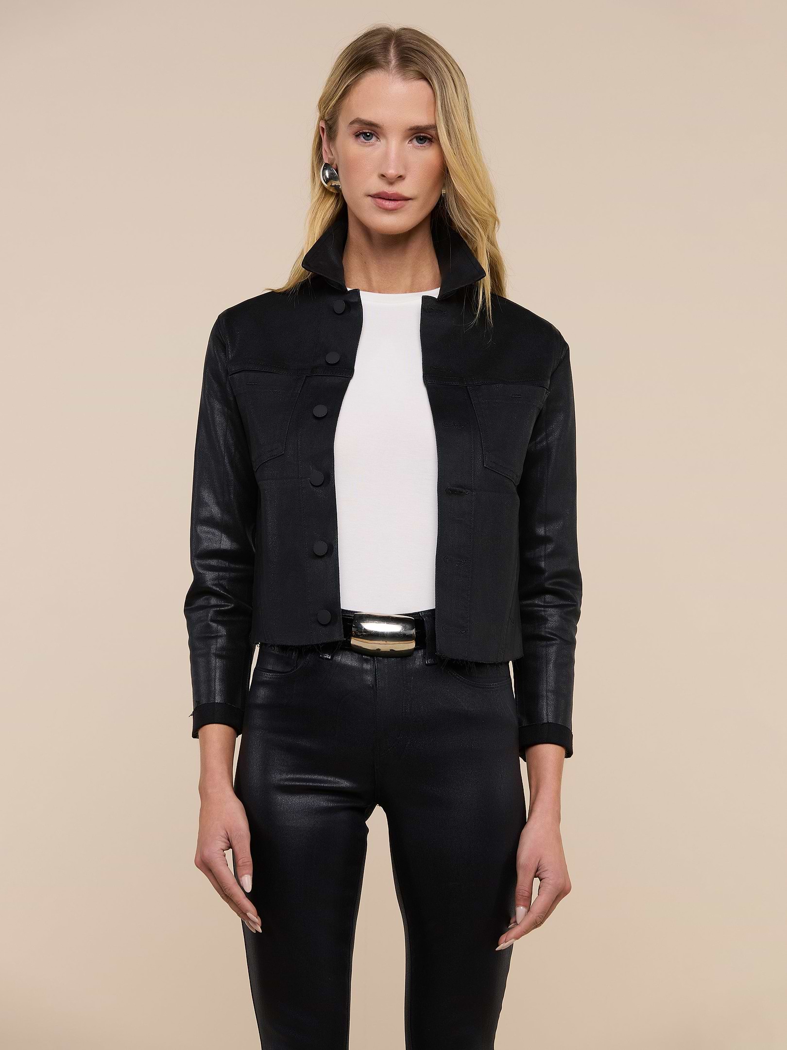 A woman stands facing forward, wearing the Janelle Coated Jean Jacket in black over a white top, paired with black pants and a belt. She has shoulder-length blonde hair styled in a crop cut and is posing against a plain light background.