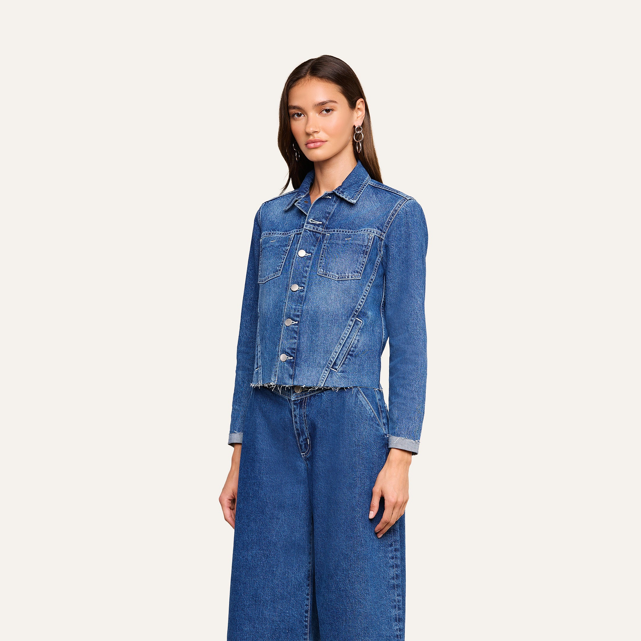 A woman stands facing forward, wearing the Janelle Jean Jacket in a medium wash with front pockets and a slim fit, paired with matching wide-leg blue jeans against a plain light background.
