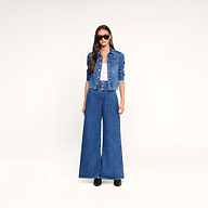 A woman wearing sunglasses, the Janelle Jean Jacket in a medium wash, a white top, and wide-leg blue jeans stands against a plain white background.