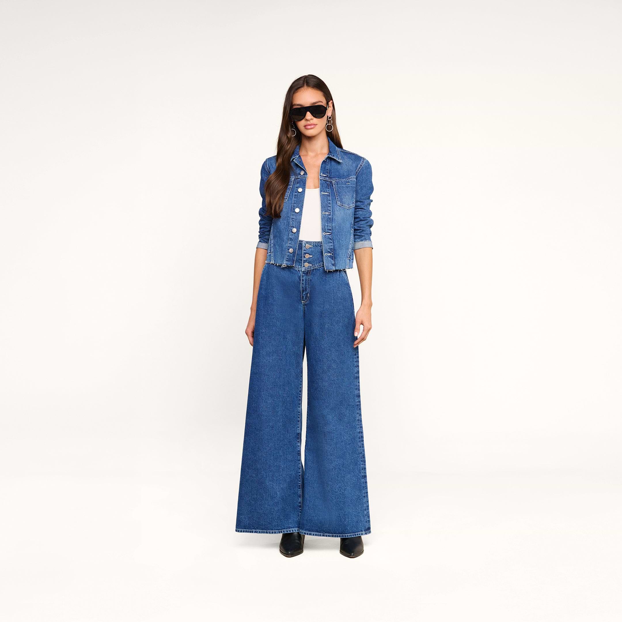A woman wearing sunglasses, the Janelle Jean Jacket in a medium wash, a white top, and wide-leg blue jeans stands against a plain white background.
