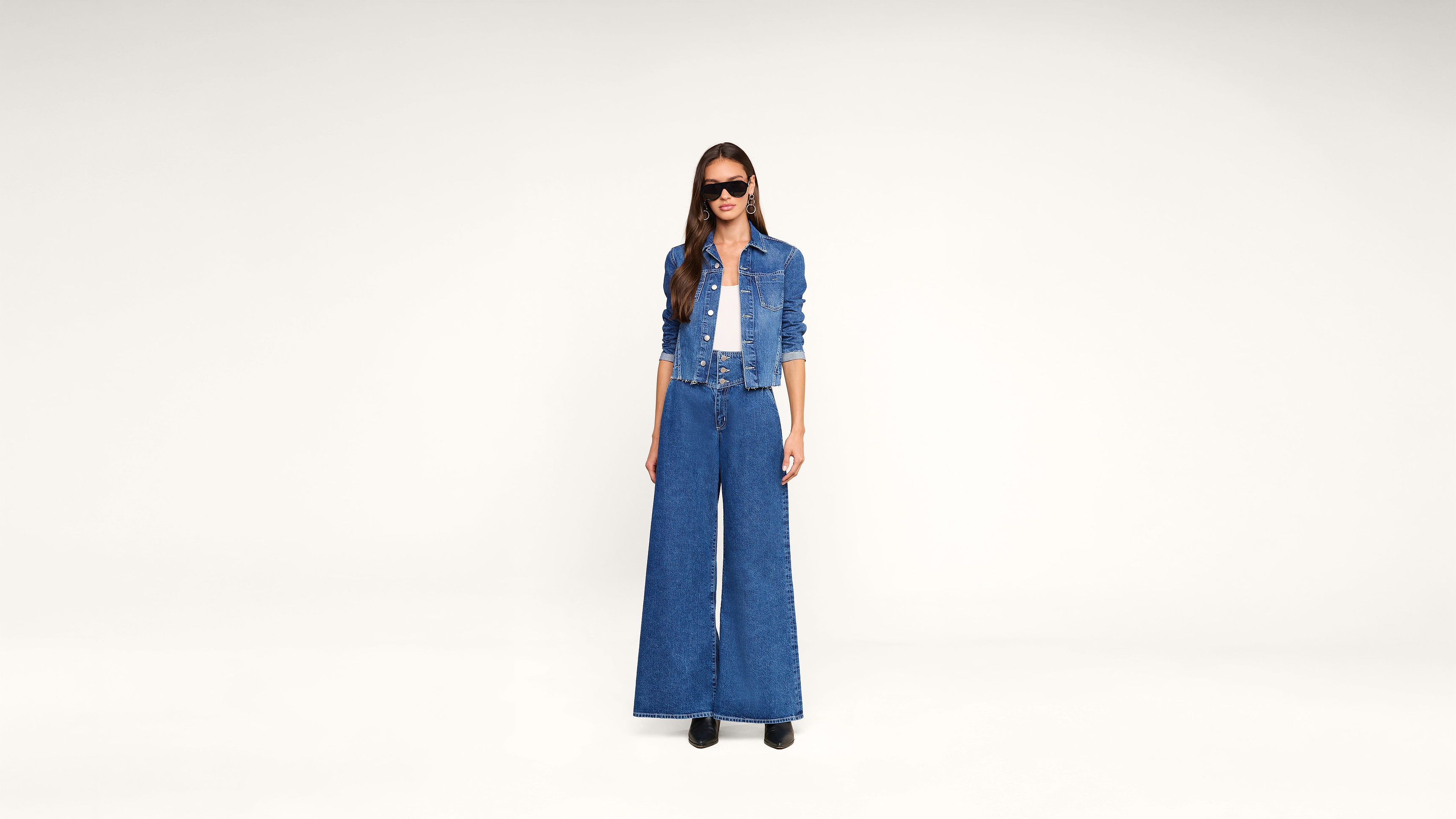 A woman stands against a plain background wearing a cropped denim jacket, wide-leg high-waisted jeans, a white top, black shoes, and dark sunglasses.