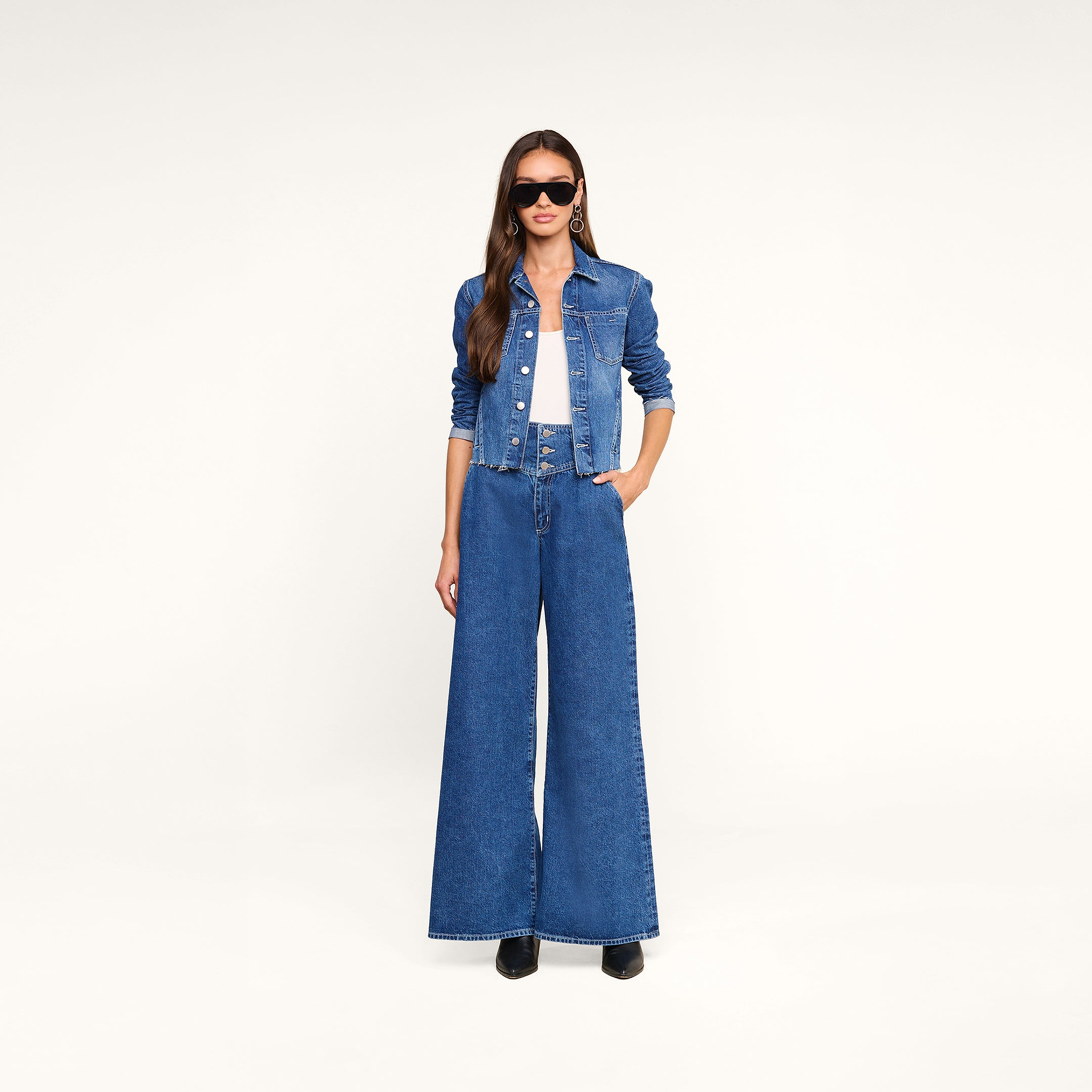 Woman wearing sunglasses, a white top, wide-leg high-waisted jeans, and the Janelle Jean Jacket stands against a plain white background with one hand in her pocket.