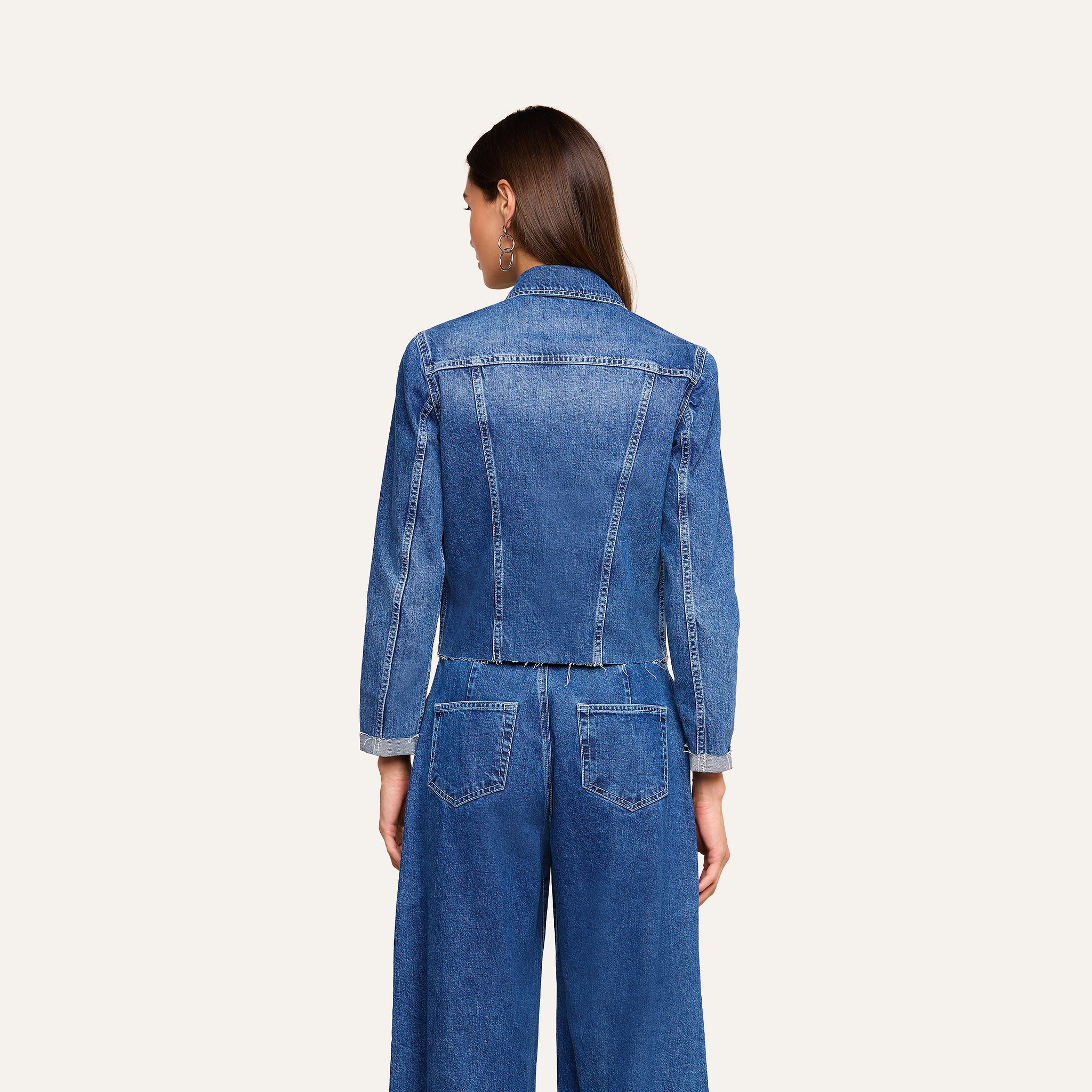 A woman with long brown hair, wearing the Janelle Jean Jacket in a medium wash and wide-leg jeans, is standing with her back to the camera against a plain, light background.
