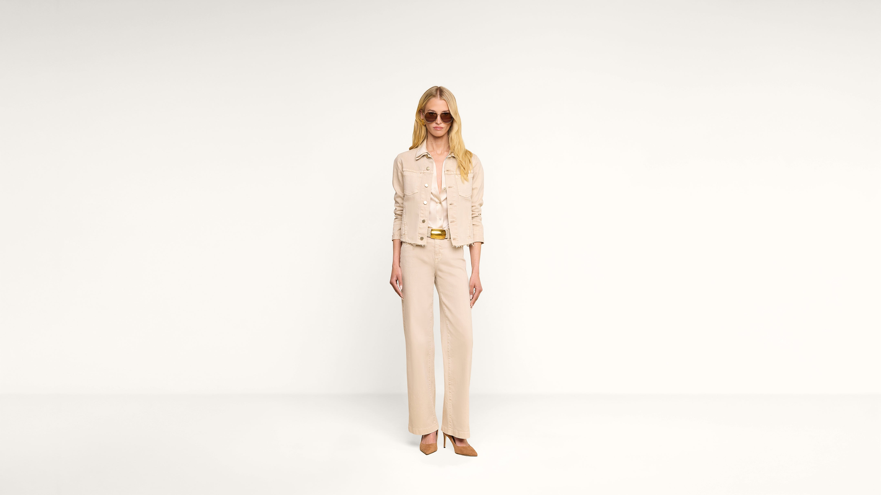 A woman wearing sunglasses and an all-beige outfit, including a jacket and wide-leg pants, stands against a plain white background.