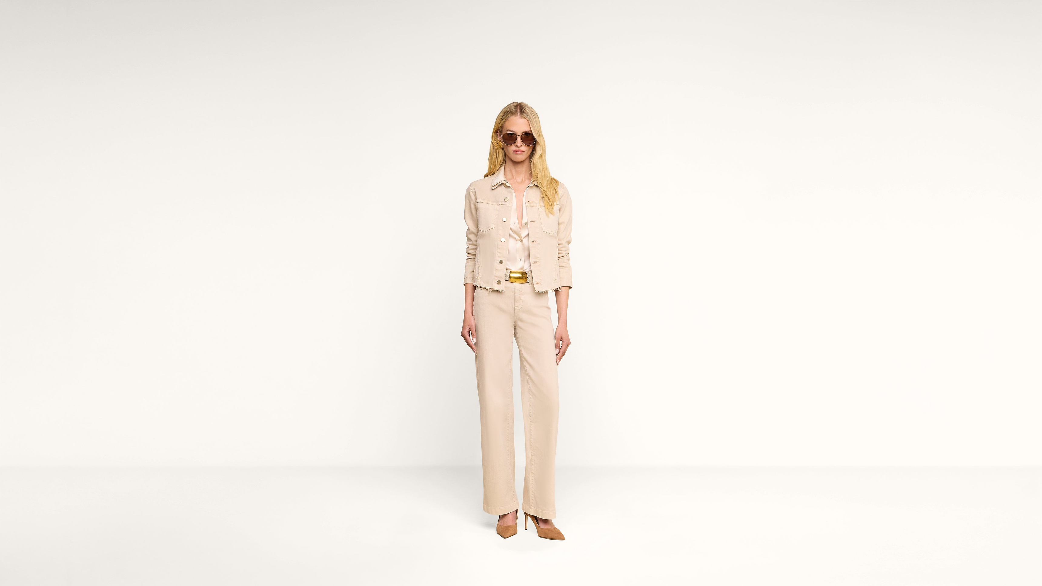 A woman wearing sunglasses and an all-beige outfit, including a jacket and wide-leg pants, stands against a plain white background.