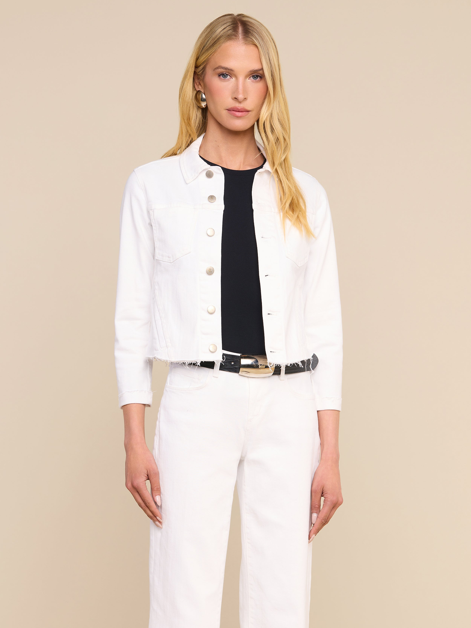 A woman with long blonde hair wears the Janelle Jean Jacket, styled with white pants over a black top, standing against a plain light background.