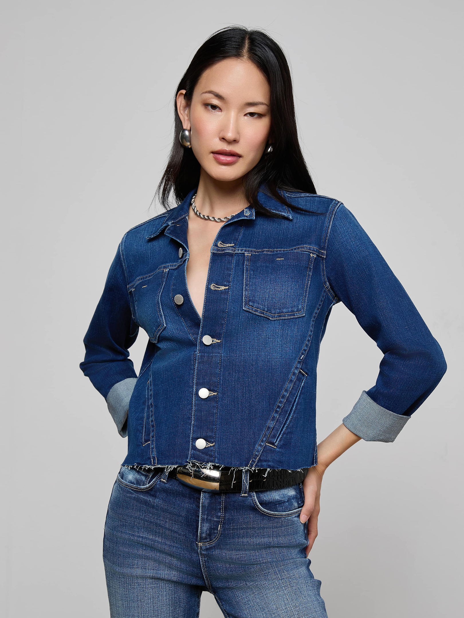 A woman stands against a plain background wearing the Janelle Jean Jacket in indigo, paired with matching jeans. With long dark hair, statement earrings, and a necklace, she places one hand on her hip, exuding confidence and style.