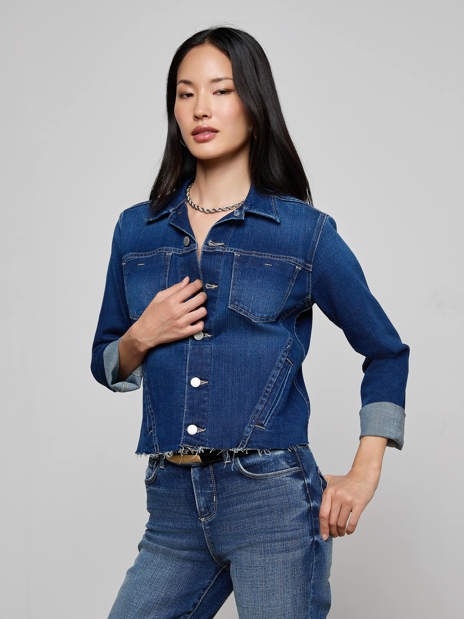 A woman wearing the Janelle Jean Jacket and matching indigo jeans poses with one hand on her waist and the other touching her jacket, set against a plain light background.