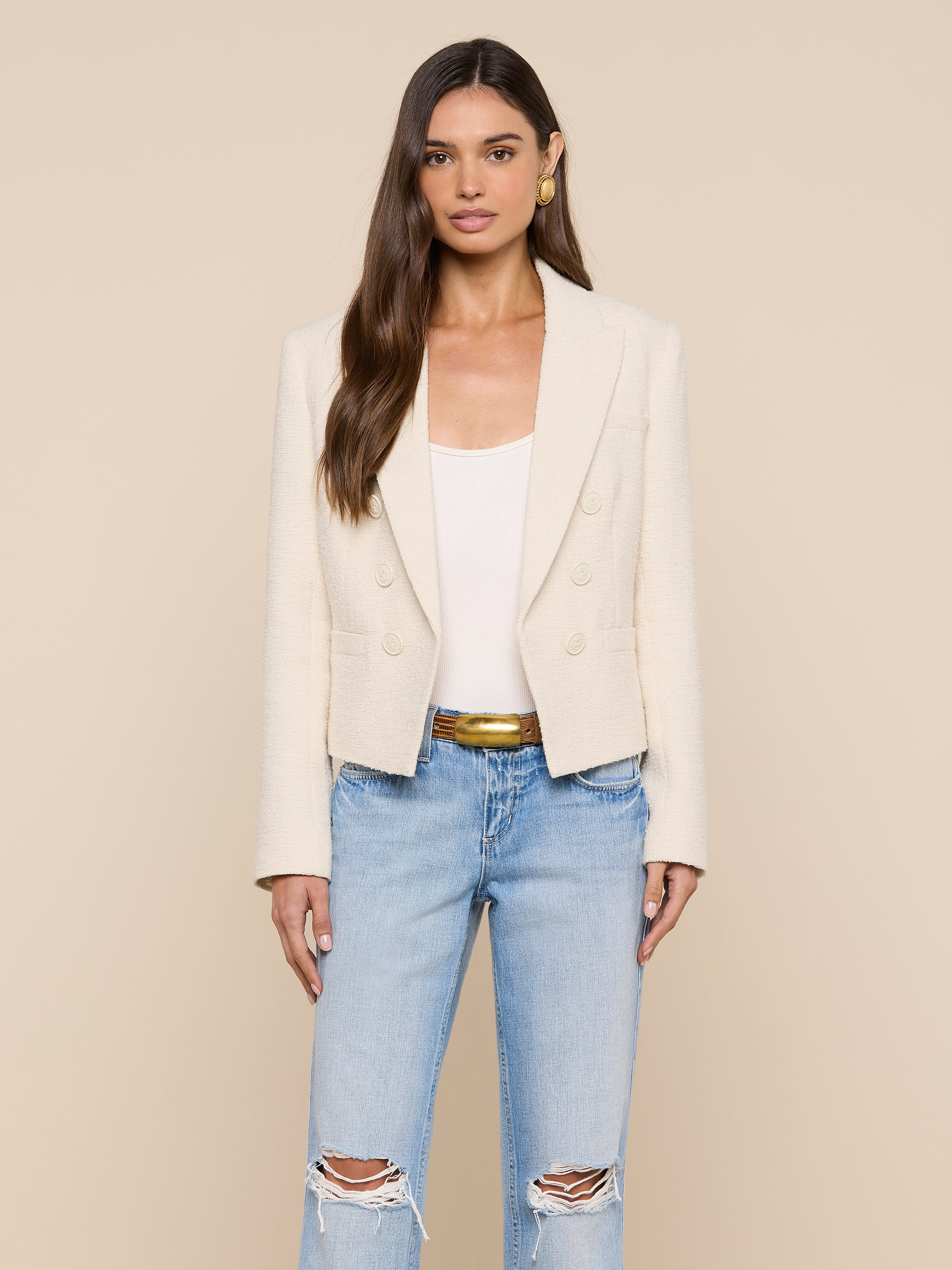 A woman stands facing forward against a plain light background, wearing the cream Brooke Tweed Blazer over a white top, paired with light blue ripped jeans and a gold belt.