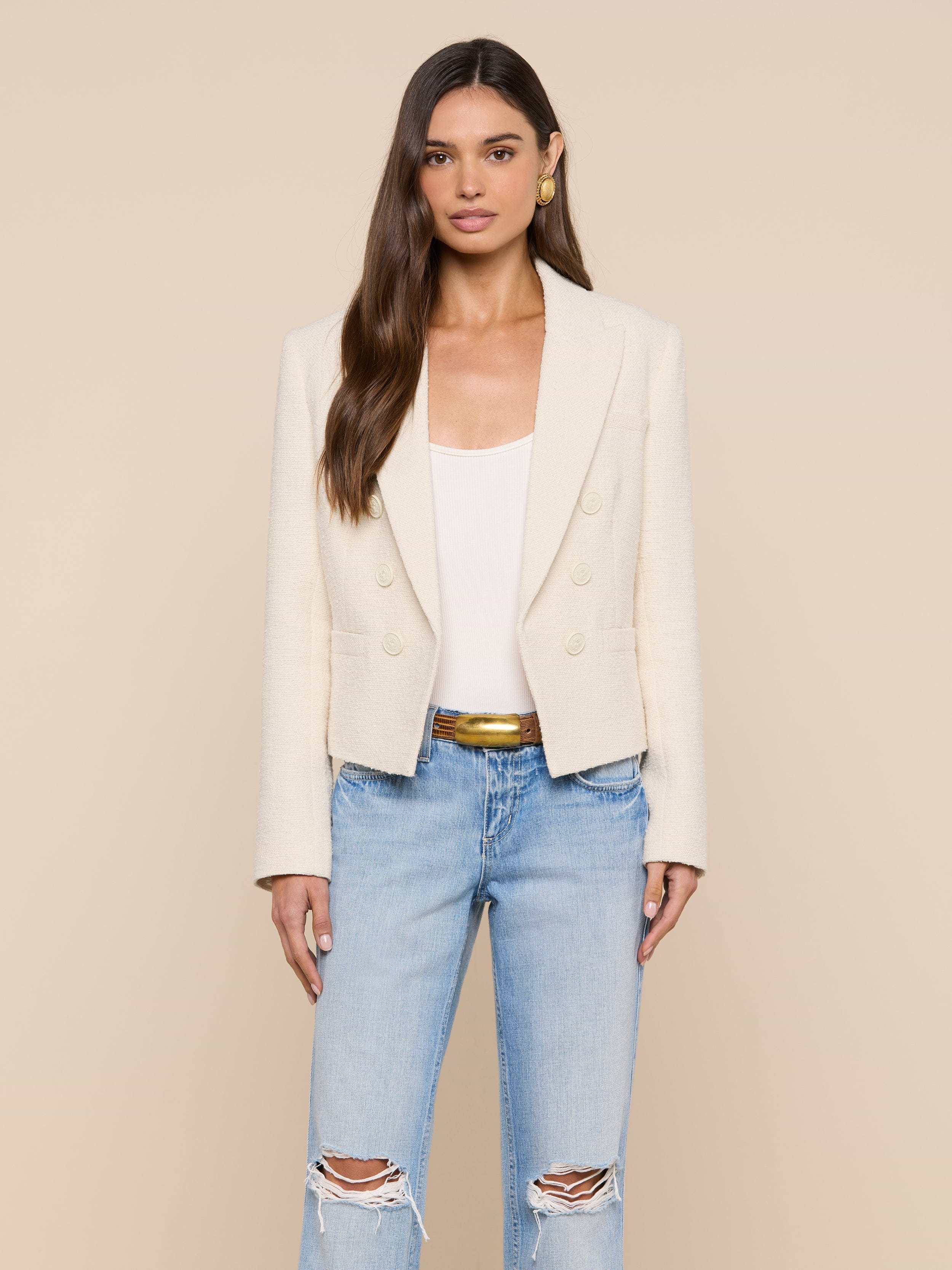 A woman stands facing forward against a plain light background, wearing the cream Brooke Tweed Blazer over a white top, paired with light blue ripped jeans and a gold belt.