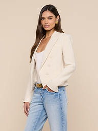 A woman with long brown hair wears the Brooke Tweed Blazer—a cream open-front style—over a white top and light blue jeans, standing against a plain light background and looking at the camera with a slight smile.