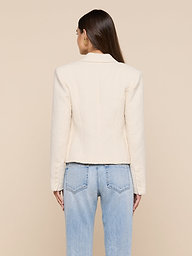 A woman stands facing away, wearing the Brooke Tweed Blazer in a cream hue paired with light blue jeans. Her straight brown hair cascades down her back, set against a plain, light-colored background.