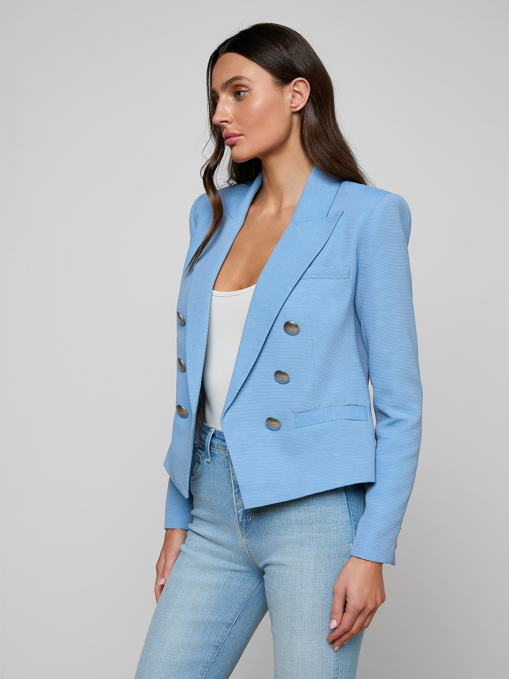 A woman wears the open-front Brooke Blazer, crafted from lightweight ribbed fabric, paired with light jeans as she stands against a plain background in a three-quarter pose facing left.