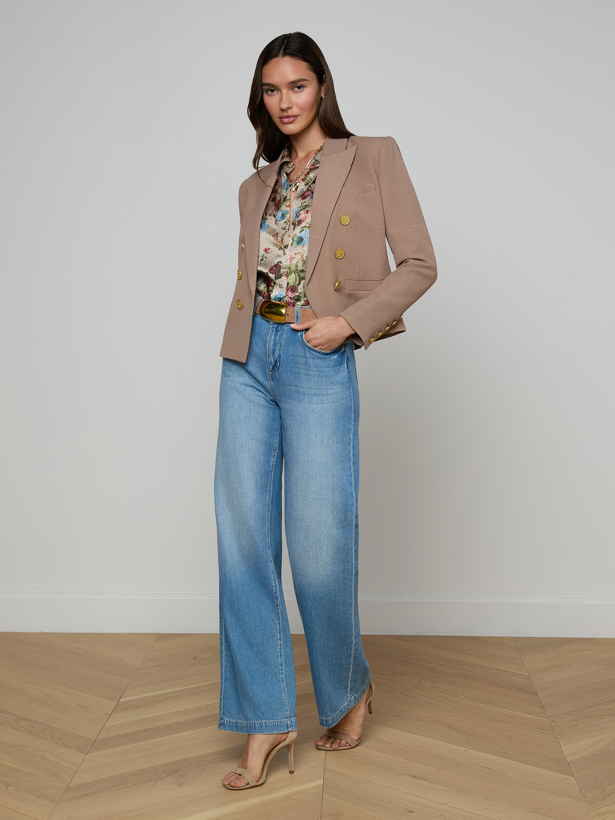 A woman stands against a plain background wearing the Brooke Blazer in tan, styled over a floral blouse with wide-leg blue jeans and beige heels, posing confidently with one hand in her pocket.