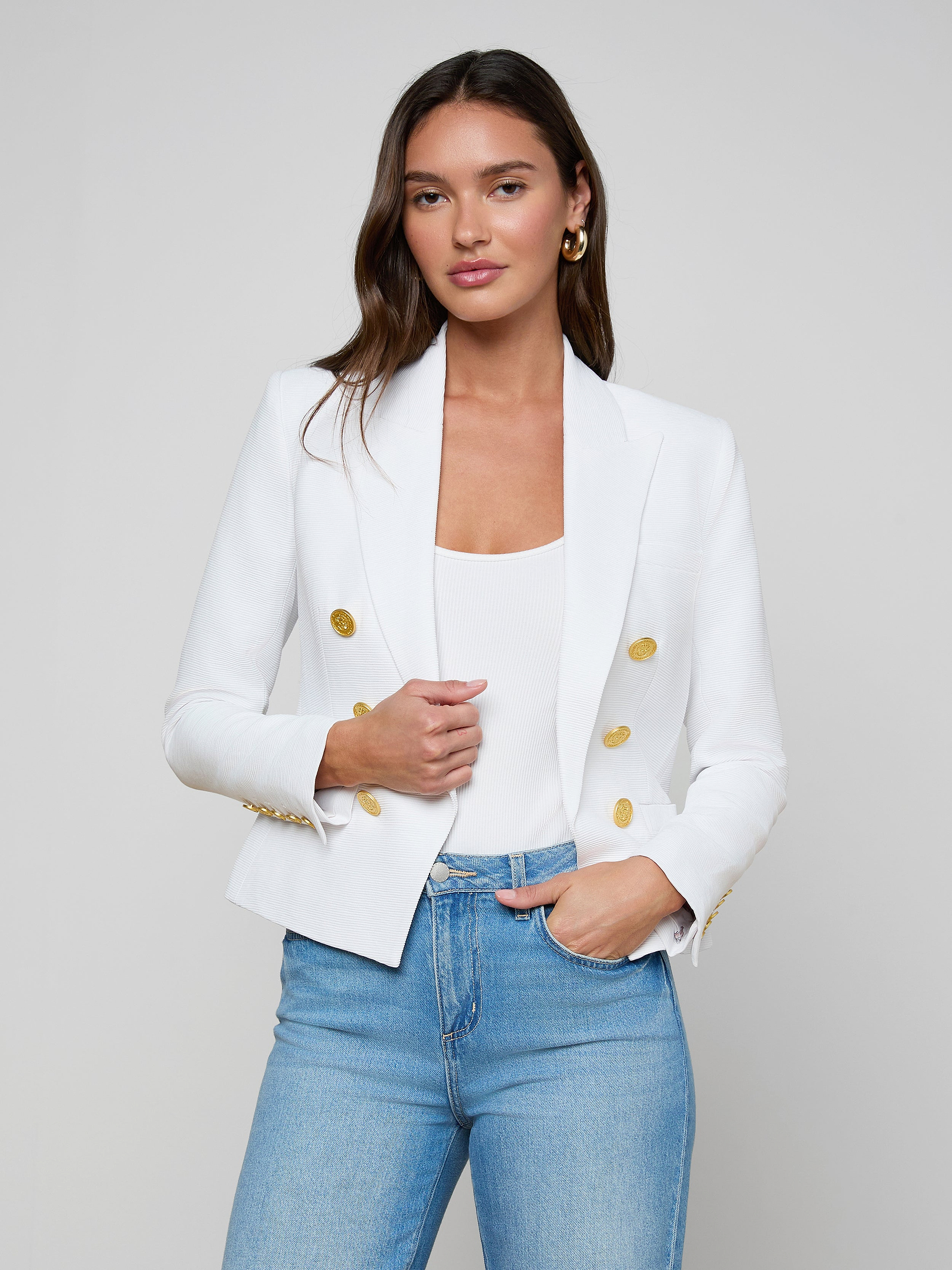 A woman stands against a plain light background with one hand in her pocket, wearing the Brooke Blazer—a white double-breasted blazer with gold buttons—paired with a white top and light blue jeans.