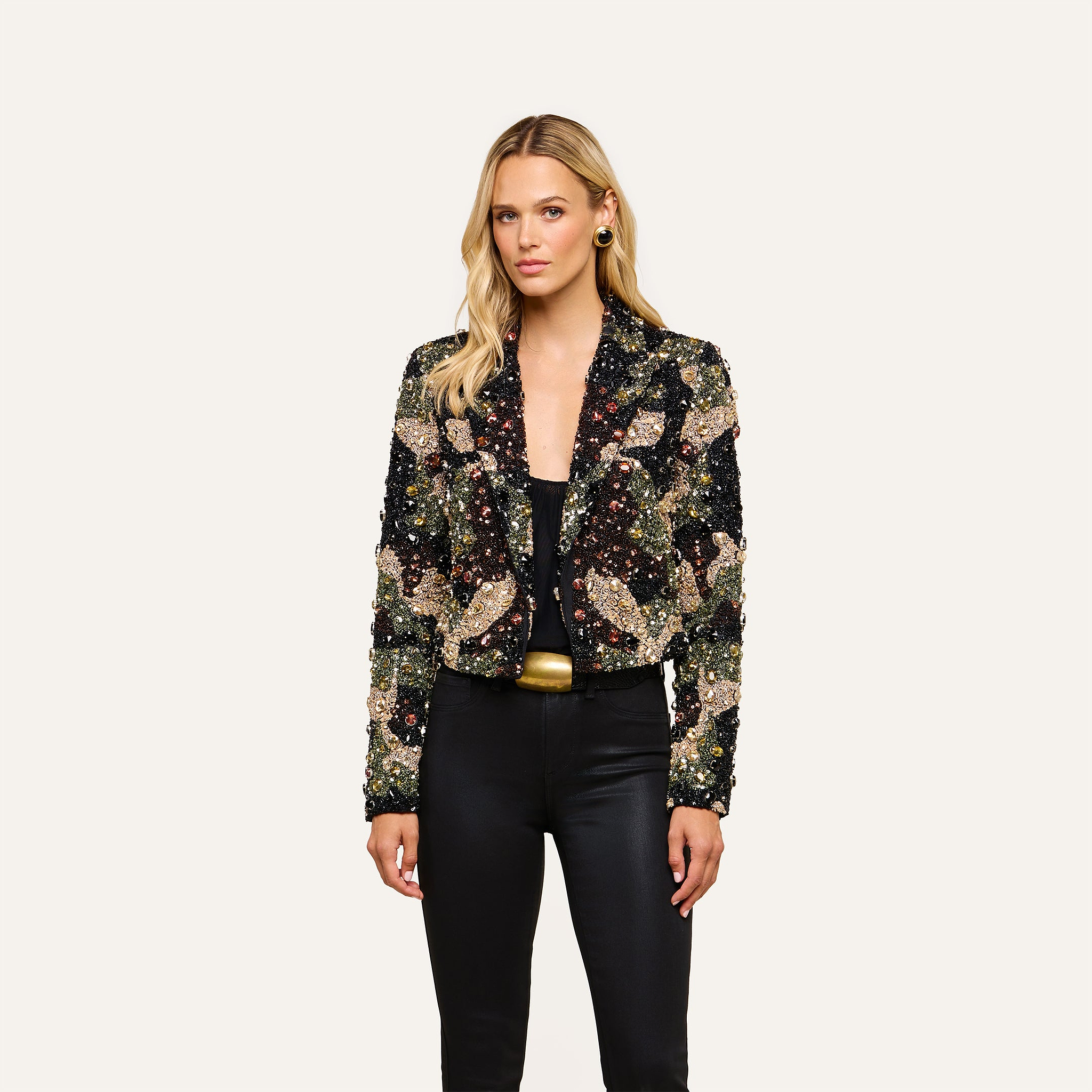 A woman wearing the Jen Crystal Blazer—a couture-inspired, hand-embellished piece—over a black top and pants stands against a plain, light background, looking at the camera with a neutral expression.