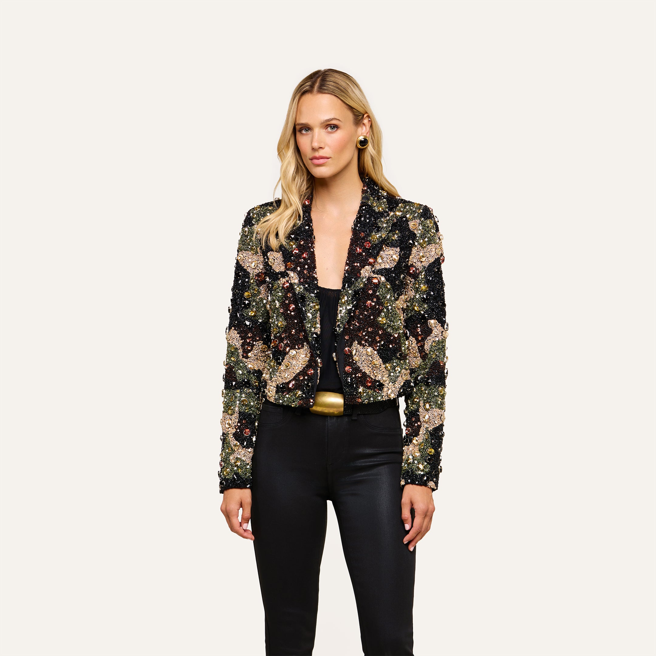 A woman wearing the Jen Crystal Blazer—a couture-inspired, hand-embellished piece—over a black top and pants stands against a plain, light background, looking at the camera with a neutral expression.