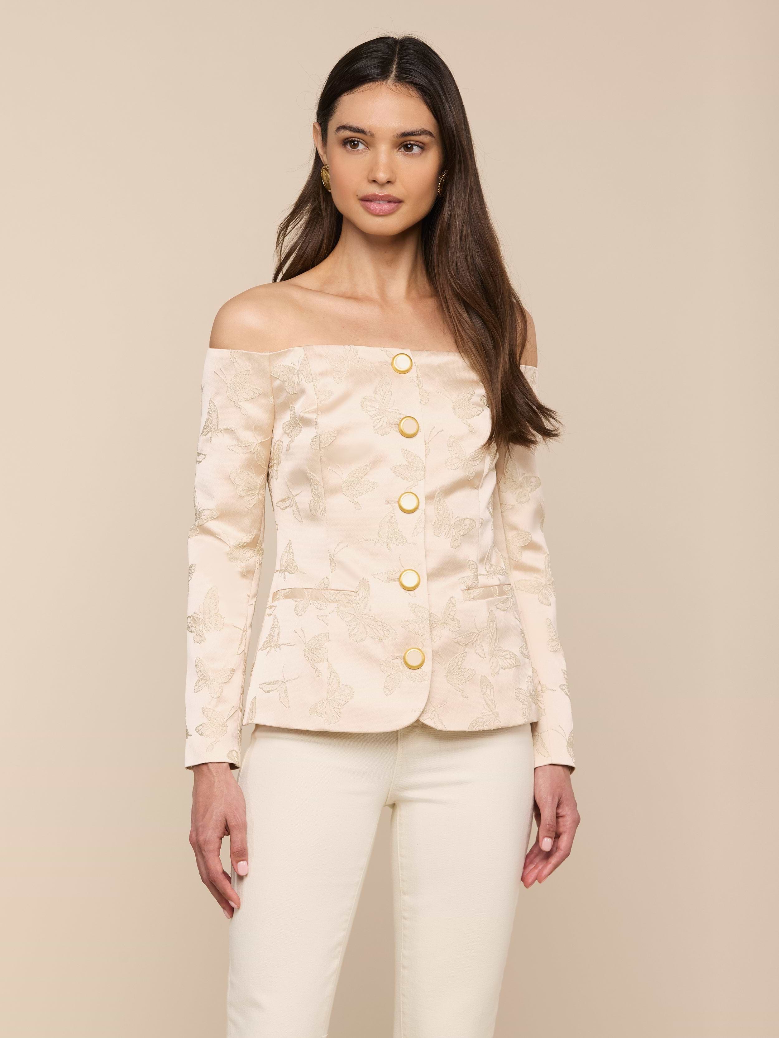 A woman stands against a plain light background, wearing the Alice Blazer Top—an off-the-shoulder cream blazer with gold buttons—paired with matching cream tailored suiting pants.