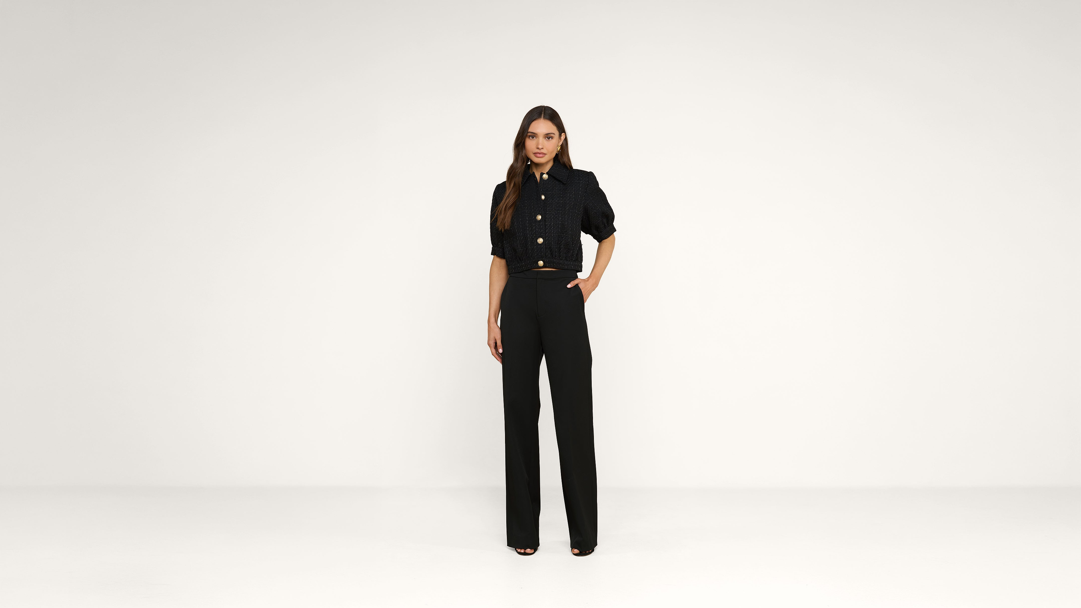 Woman stands against a plain white background, wearing a black button-up shirt and high-waisted black trousers with open-toe heels. One hand rests on her hip, and she looks straight ahead with a neutral expression.