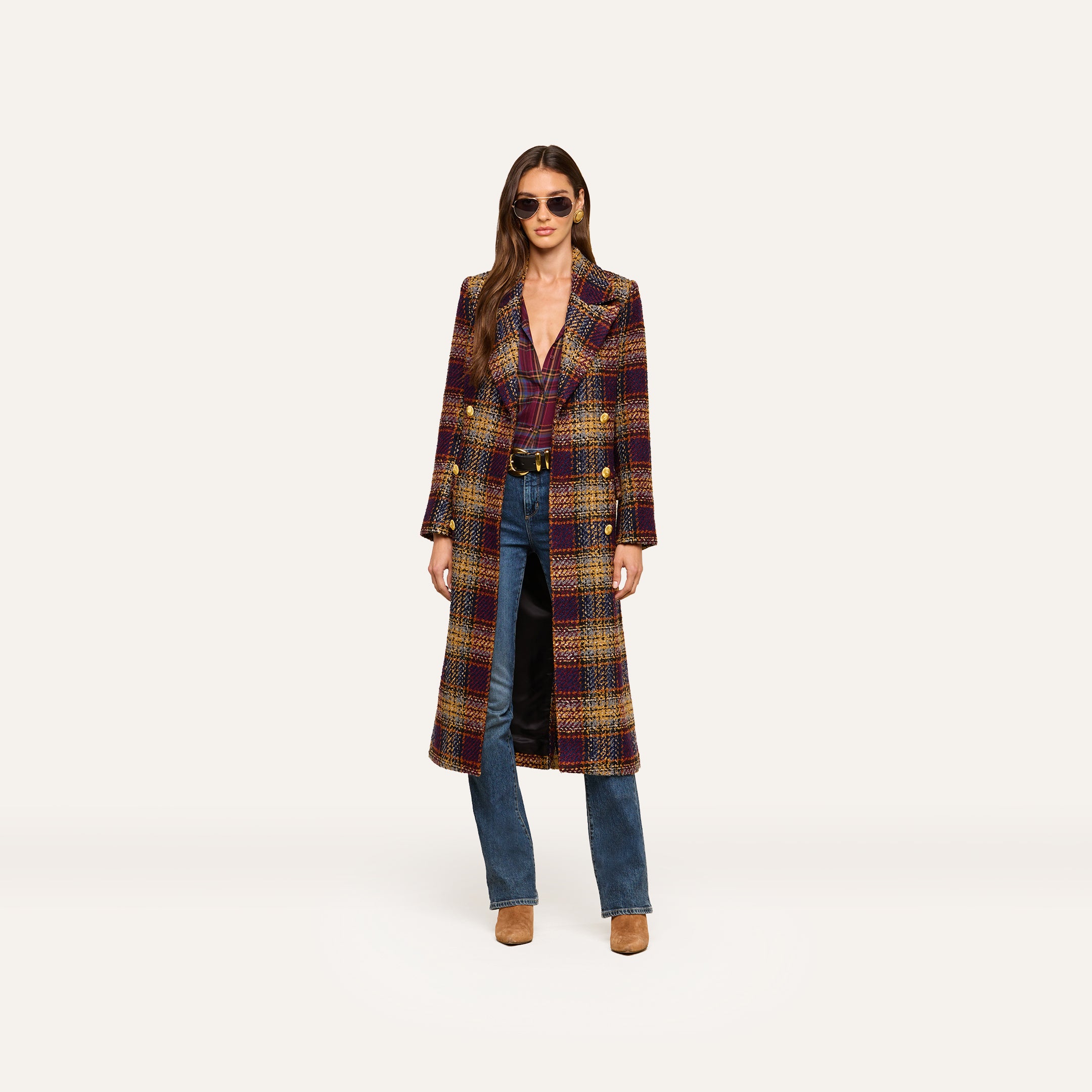 A woman wearing sunglasses and the Kolina Tweed Long Coat with gold buttons, along with blue jeans and brown shoes, stands against a plain light background.