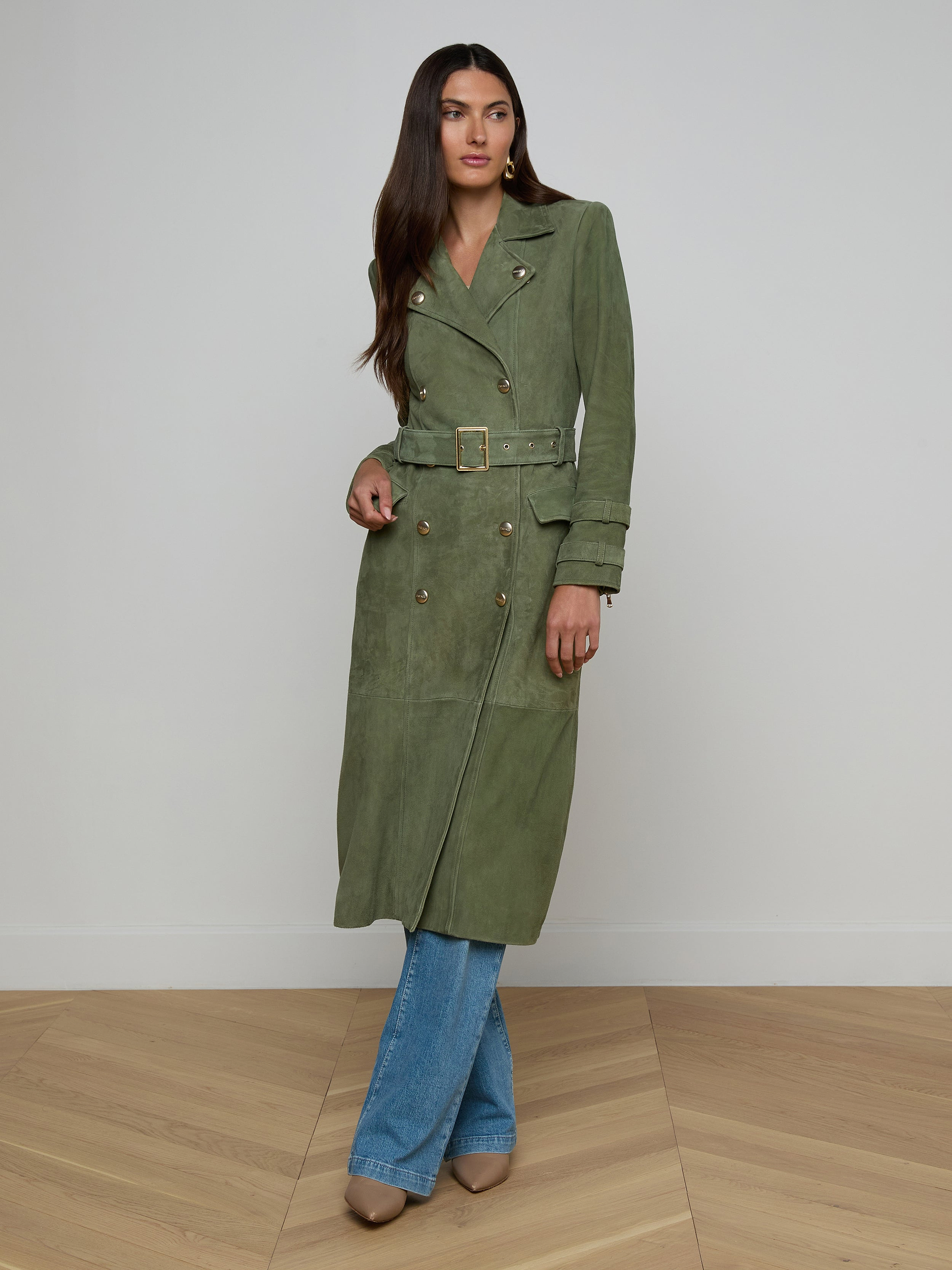 A woman models the Celina Suede Trench Coat in olive green, styled over blue jeans and beige shoes, standing against a plain white background.