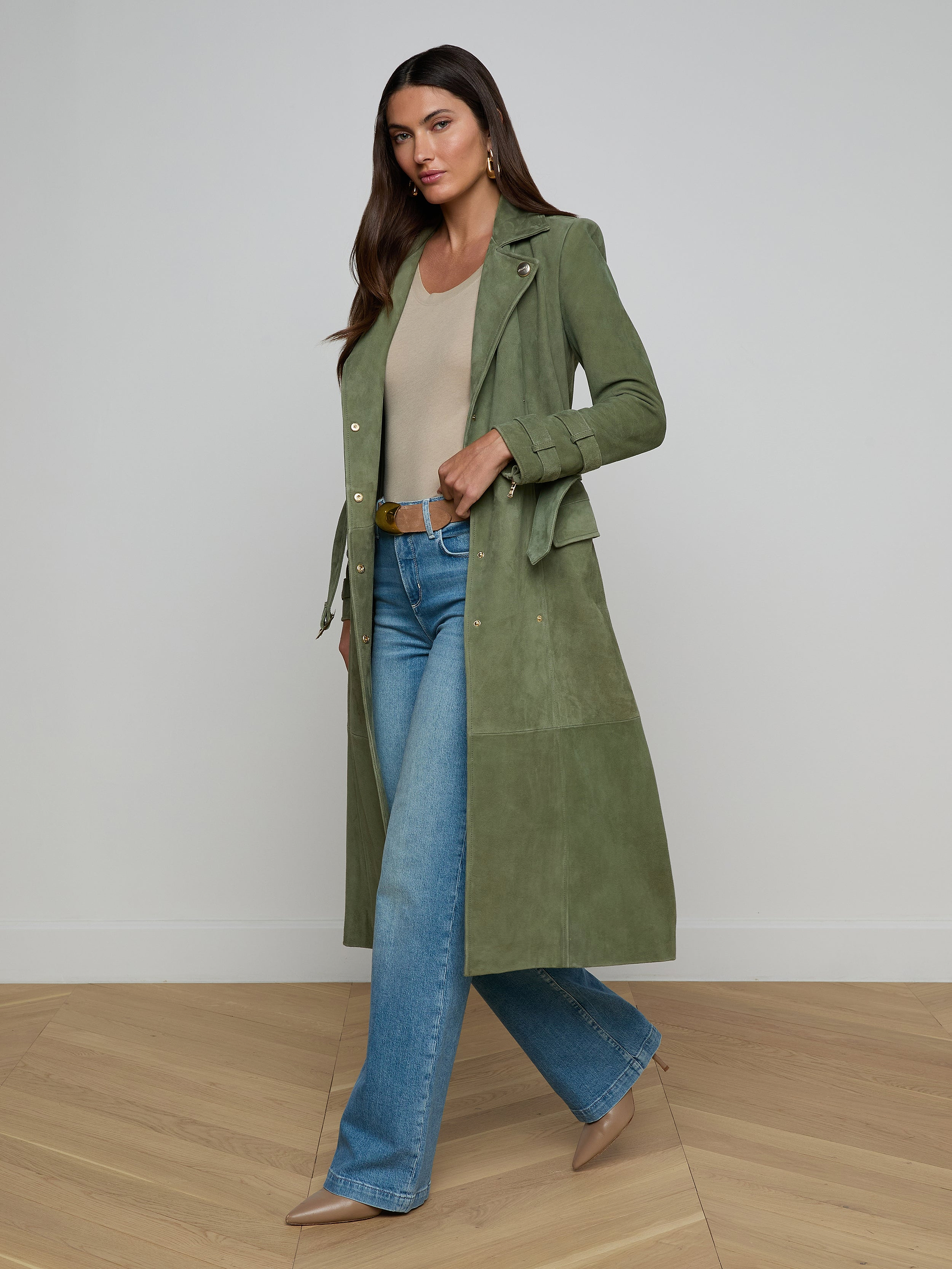 A woman wearing a long olive green Celina Suede Trench Coat, beige top, blue jeans, and tan heels walks confidently against a plain white background—showcasing a timeless wardrobe look.