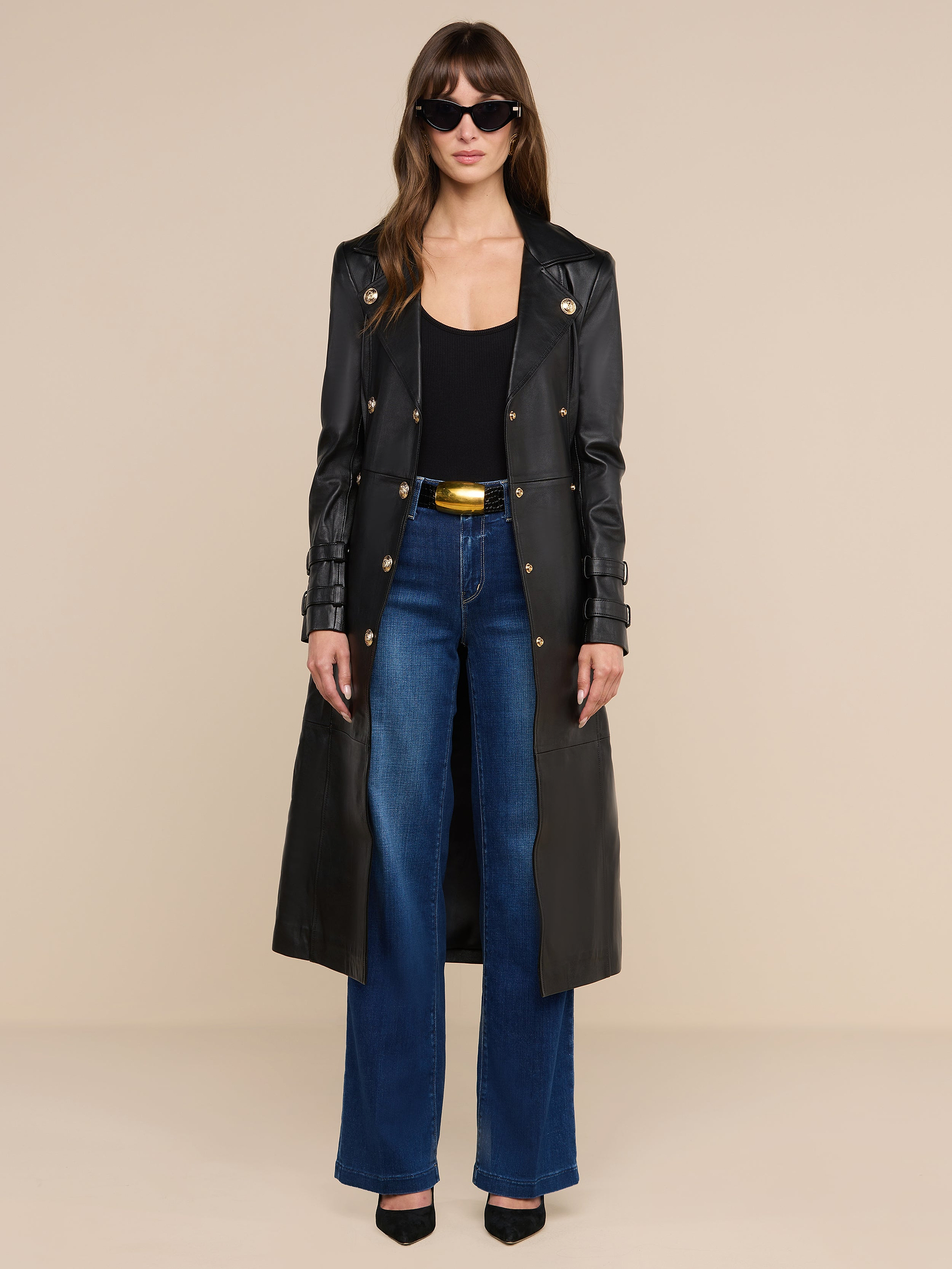 A woman stands against a plain white background wearing sunglasses, the Celina Leather Trench Coat over a black top, blue wide-leg jeans, and black heels.