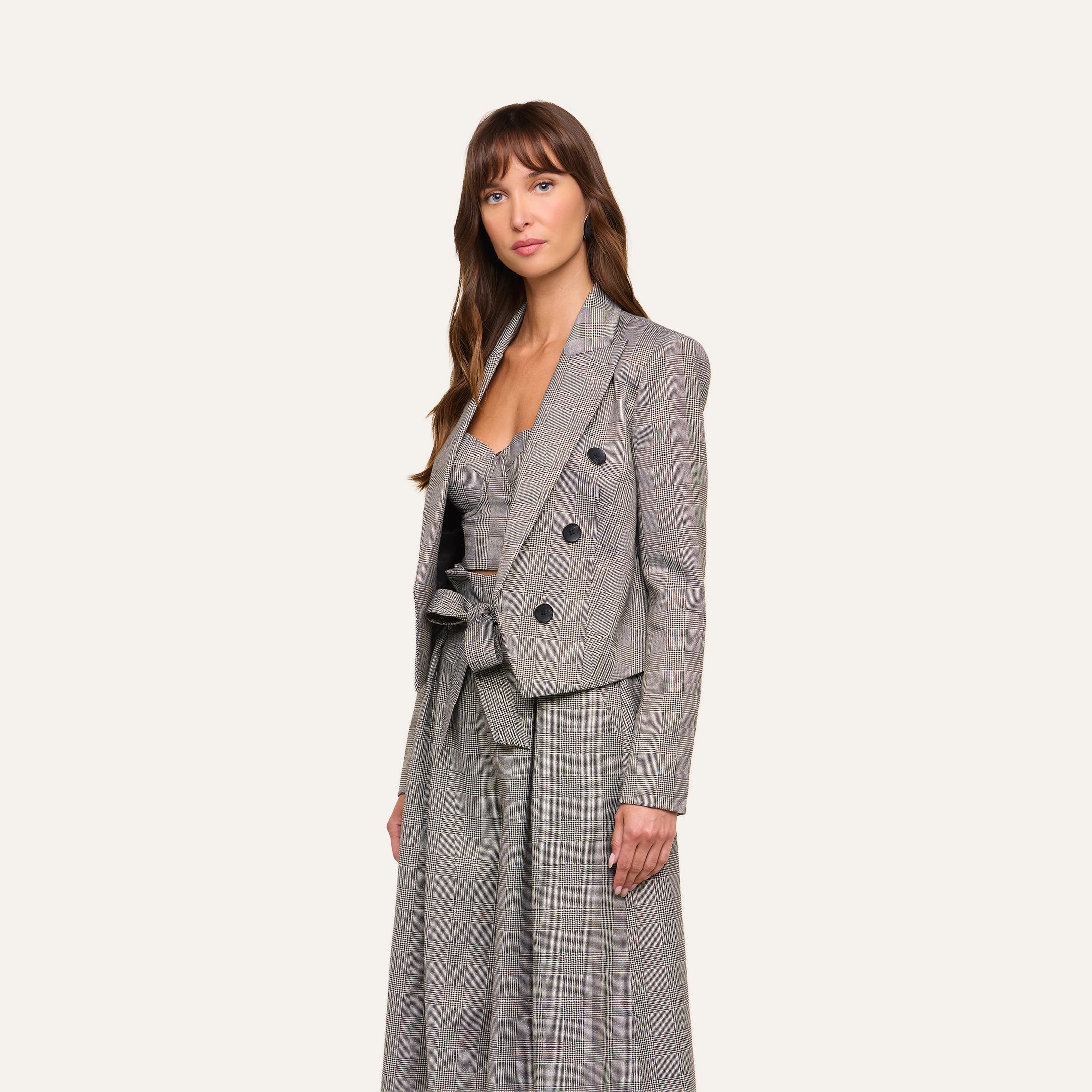 A woman wearing the Lila Tweed Blazer—a gray plaid jacket featuring a belted waist and three-button closure—stands against a plain light background.