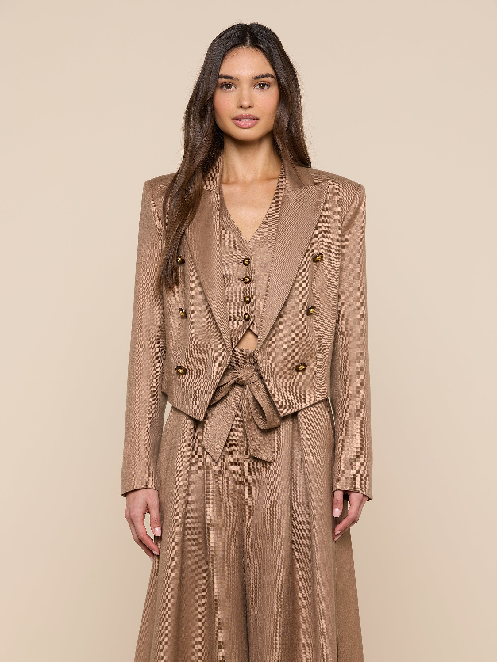 A woman stands facing forward, wearing the Lila Linen-Blend Blazer in tan with a double-breasted design, matching vest, wide-leg pants, and a tied belt. She has long, wavy brown hair and a neutral expression against a plain light background.