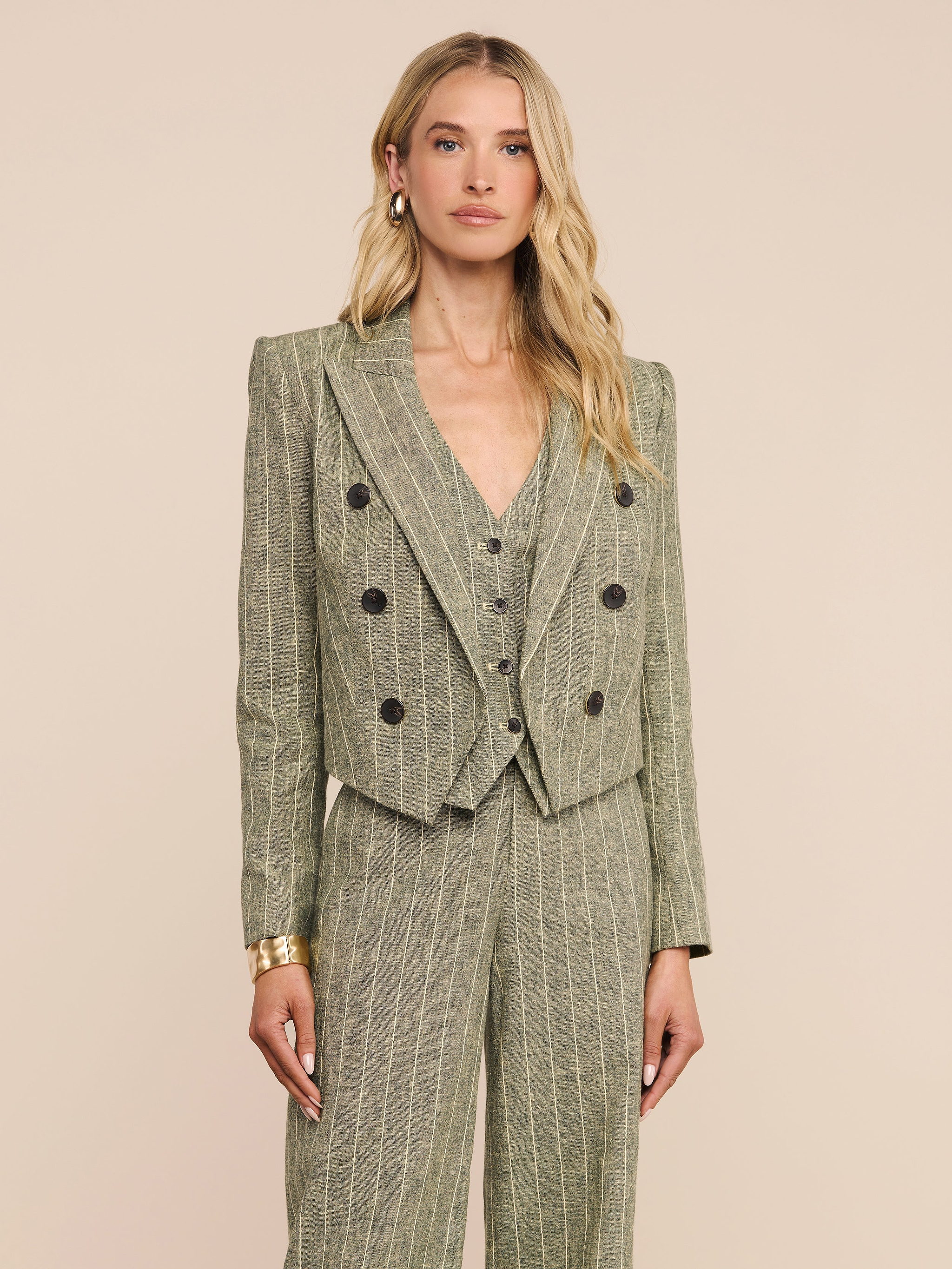 A woman with long blonde hair wears the Lila Linen-Blend Blazer—a pinstriped olive green double-breasted jacket—paired with a matching vest and wide-leg pants. She stands facing forward against a plain light background.
