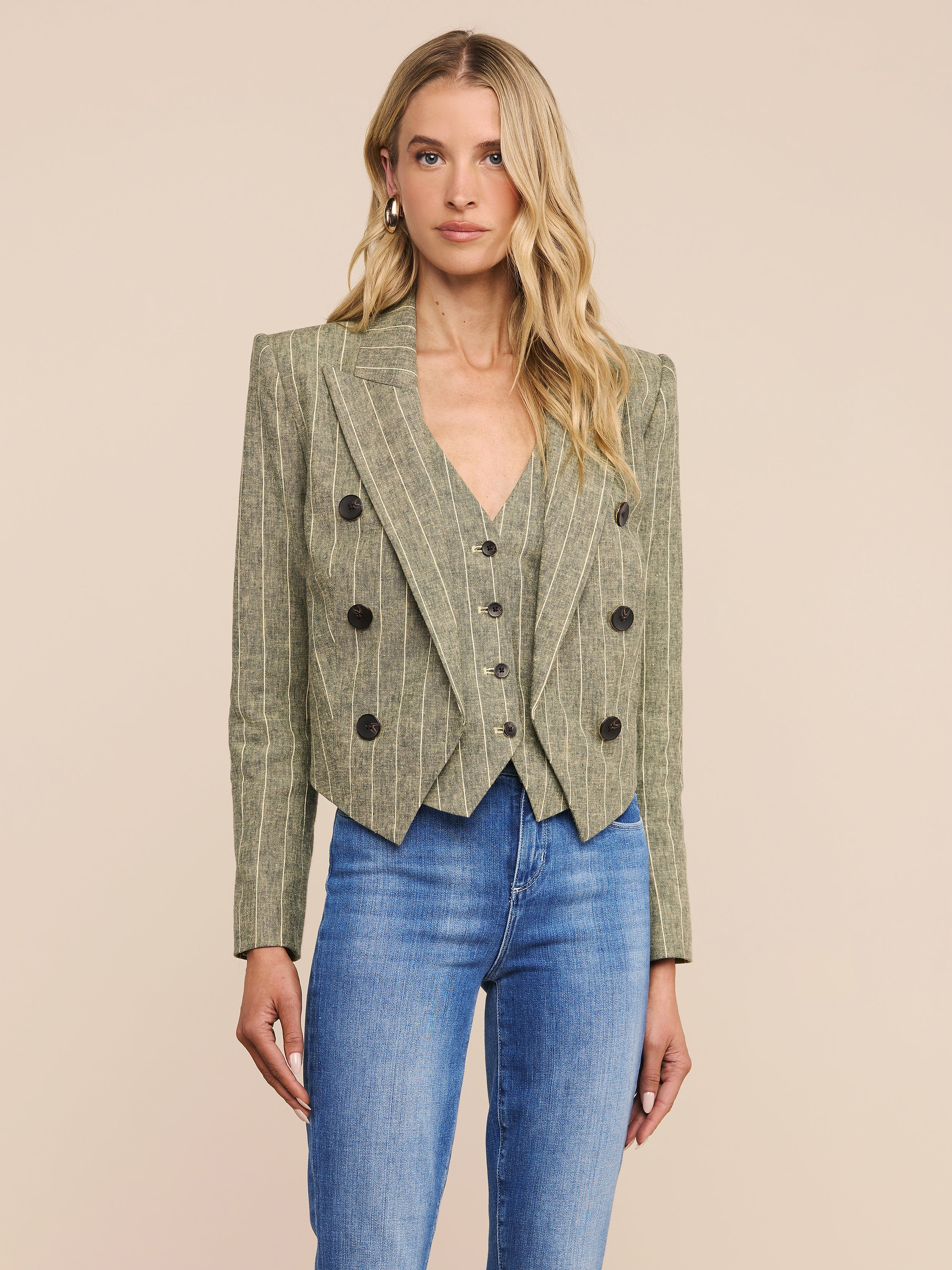 A woman with long blonde hair wears the fitted Lila Linen-Blend Blazer in pinstriped olive green, paired with blue jeans, standing facing forward against a plain light background.