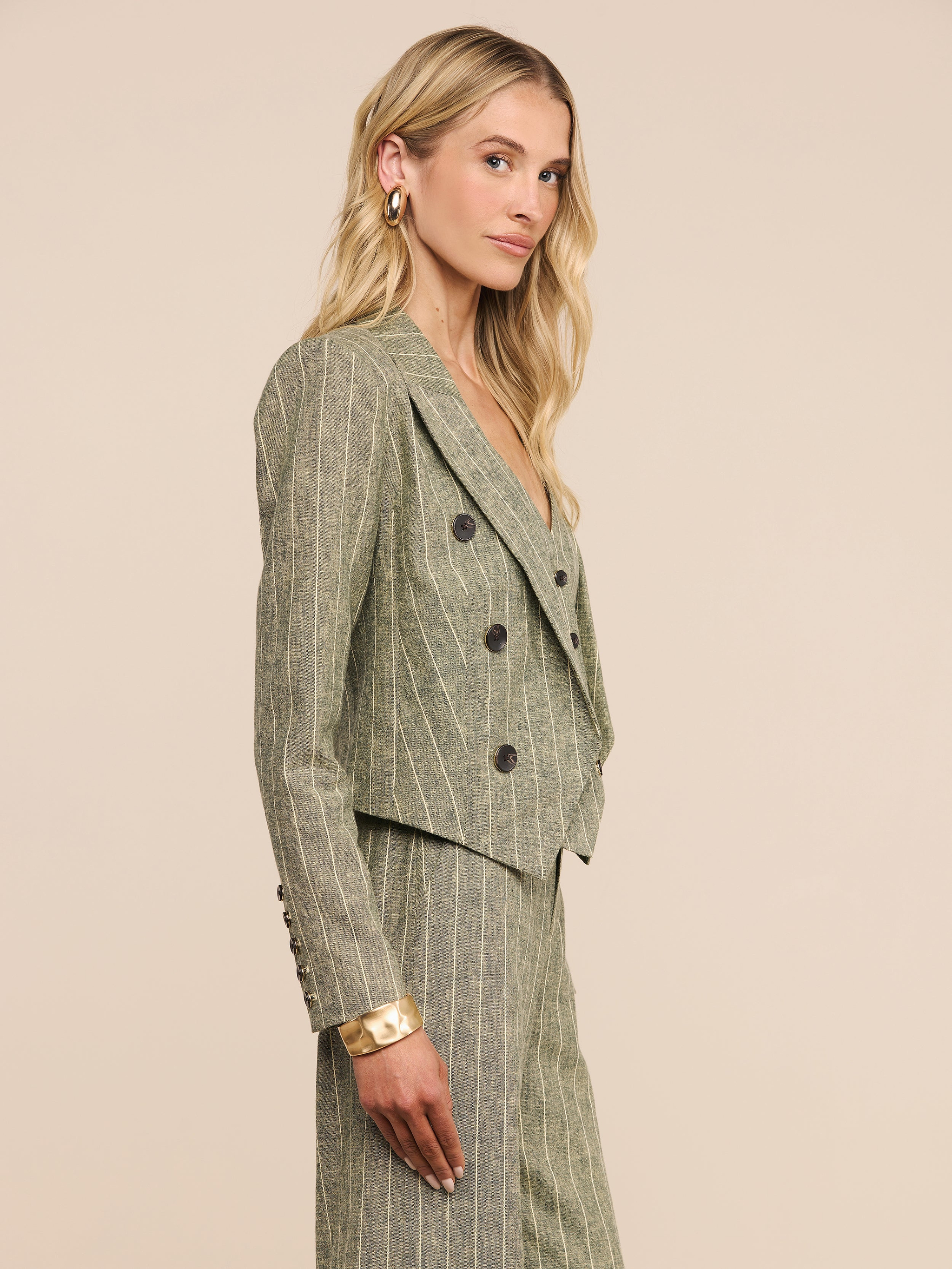 A woman with long blonde hair stands sideways against a plain white background, wearing the Lila Linen-Blend Blazer in olive green with matching pinstriped trousers. She accessorizes her look with hoop earrings and a chunky bracelet.