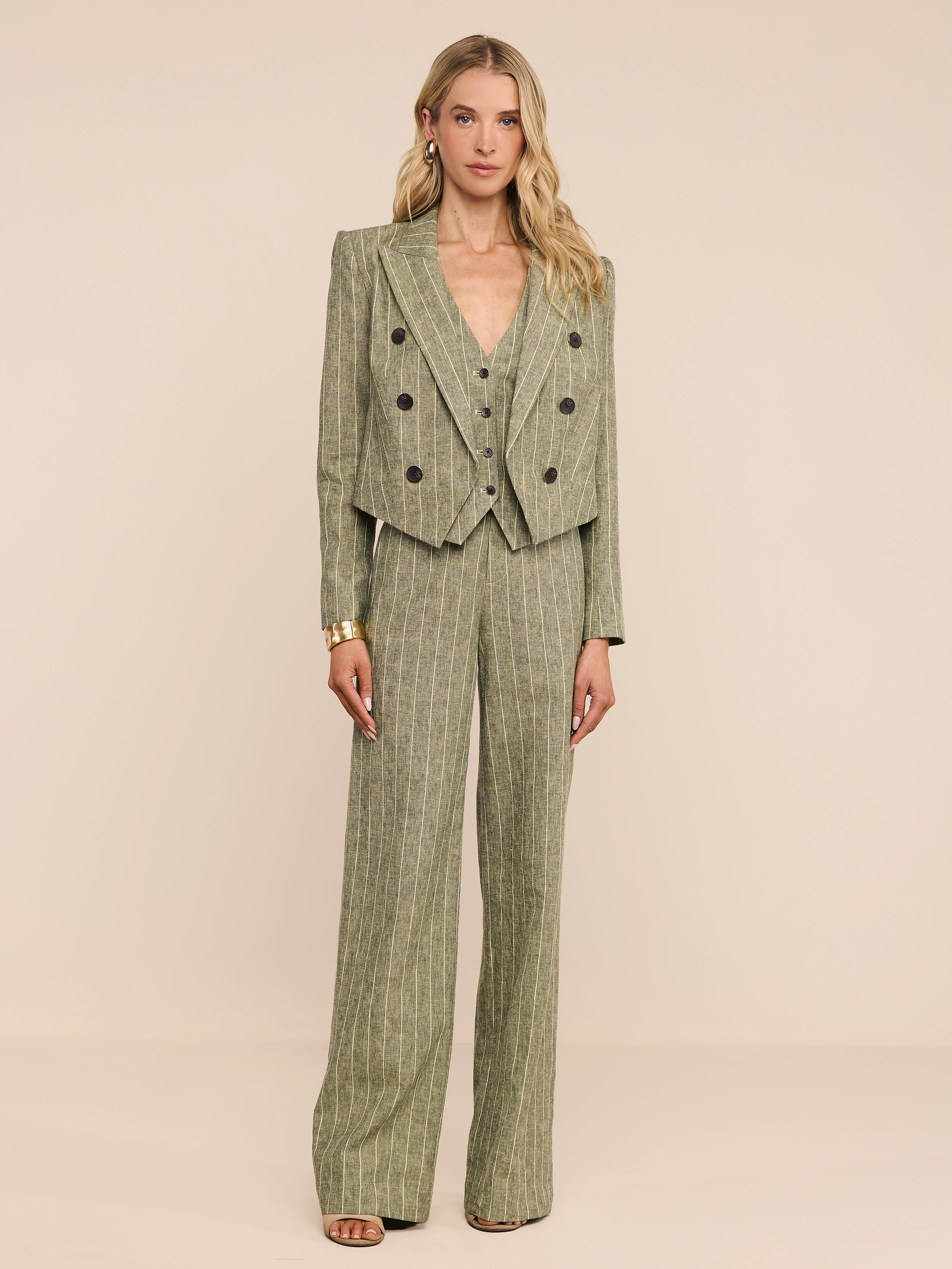 A woman stands against a plain white background wearing the Lila Linen-Blend Blazer, a cropped pinstriped olive green style paired with wide-leg trousers. She has loose blonde hair and neutral-toned heels.
