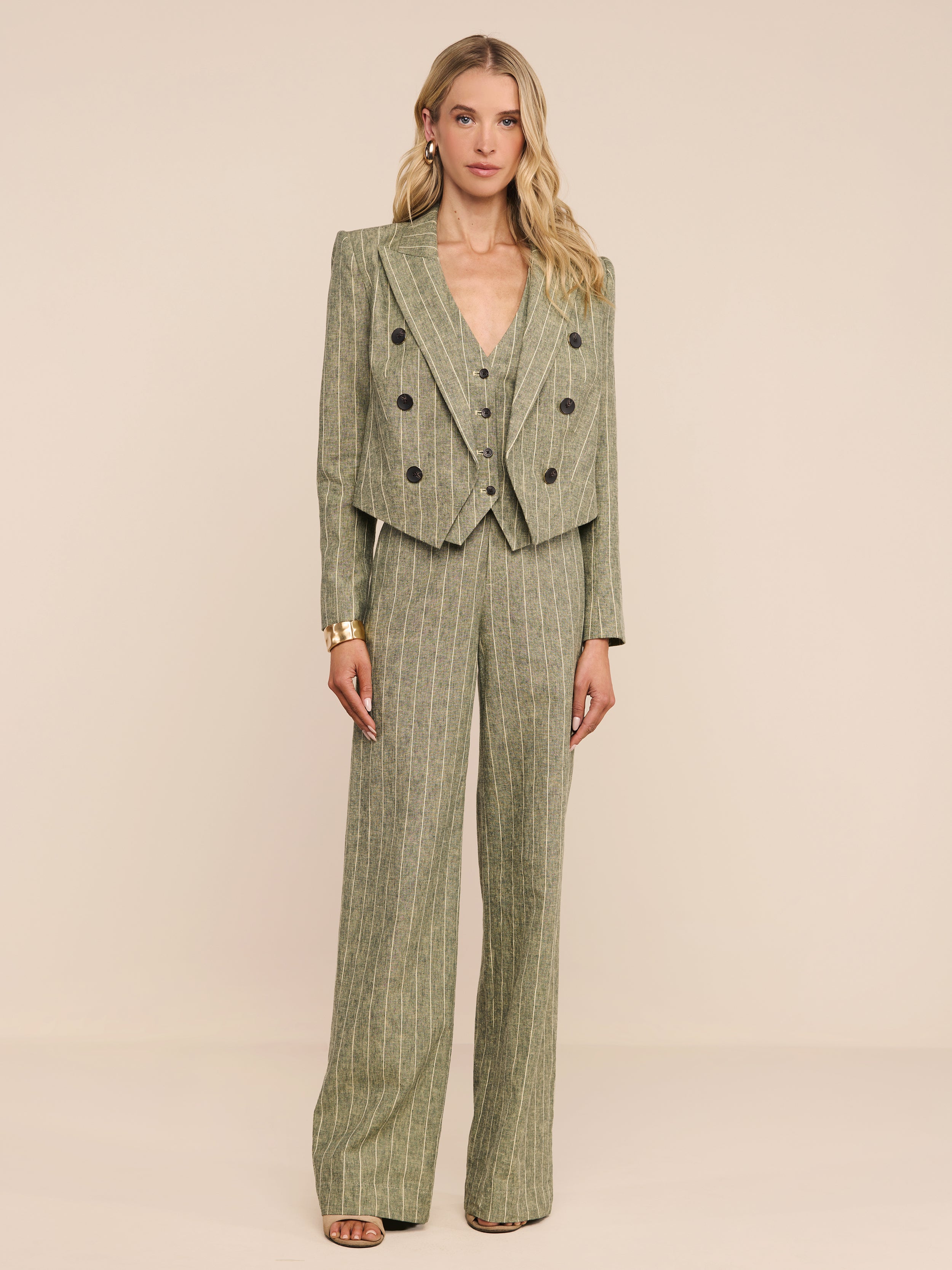 A woman stands against a plain white background wearing the Lila Linen-Blend Blazer, a cropped pinstriped olive green style paired with wide-leg trousers. She has loose blonde hair and neutral-toned heels.