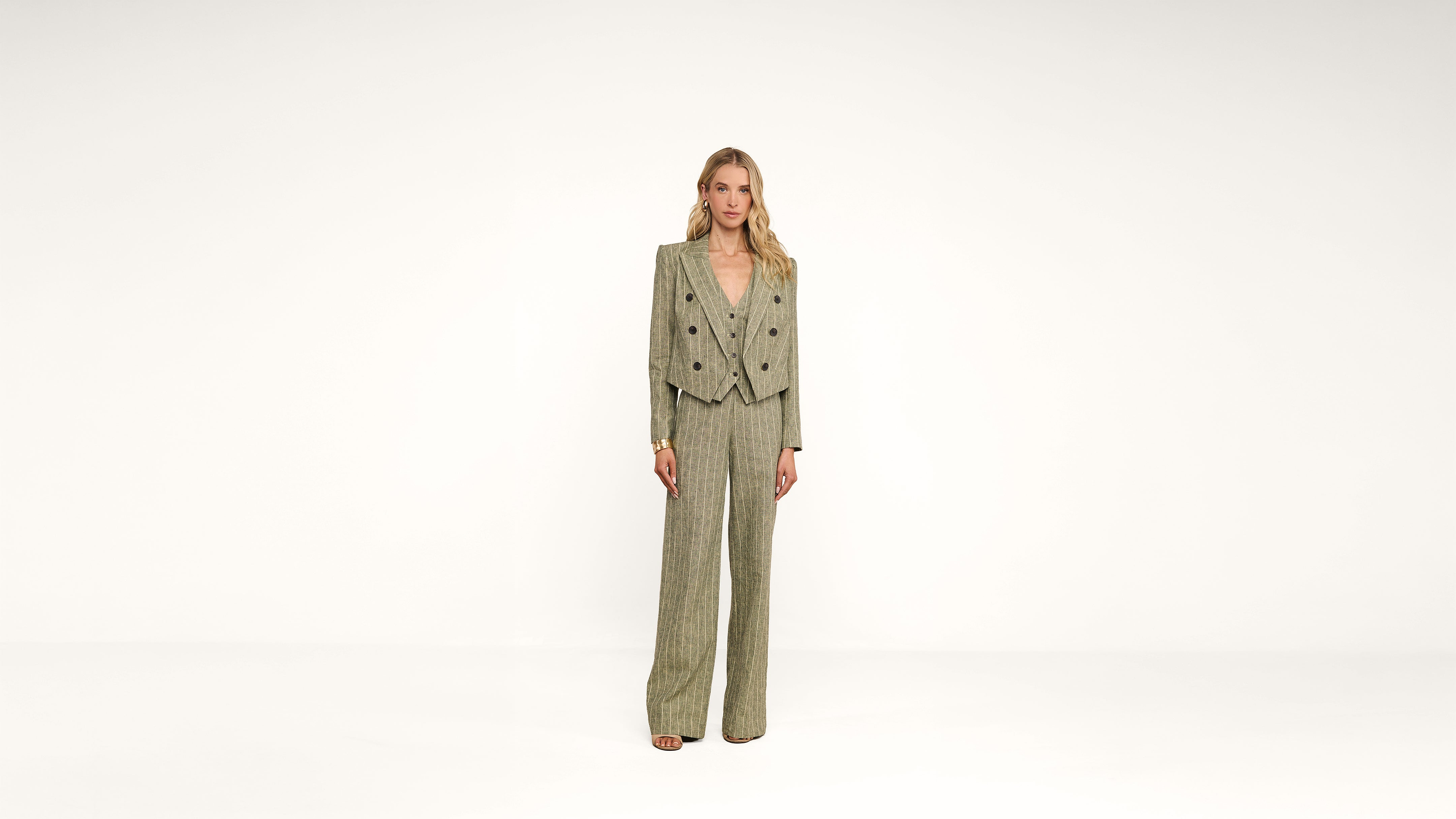 Woman stands in a light gray textured suit with wide-leg pants and a double-breasted blazer in a plain, white studio setting. She is facing forward and wearing open-toe heels.