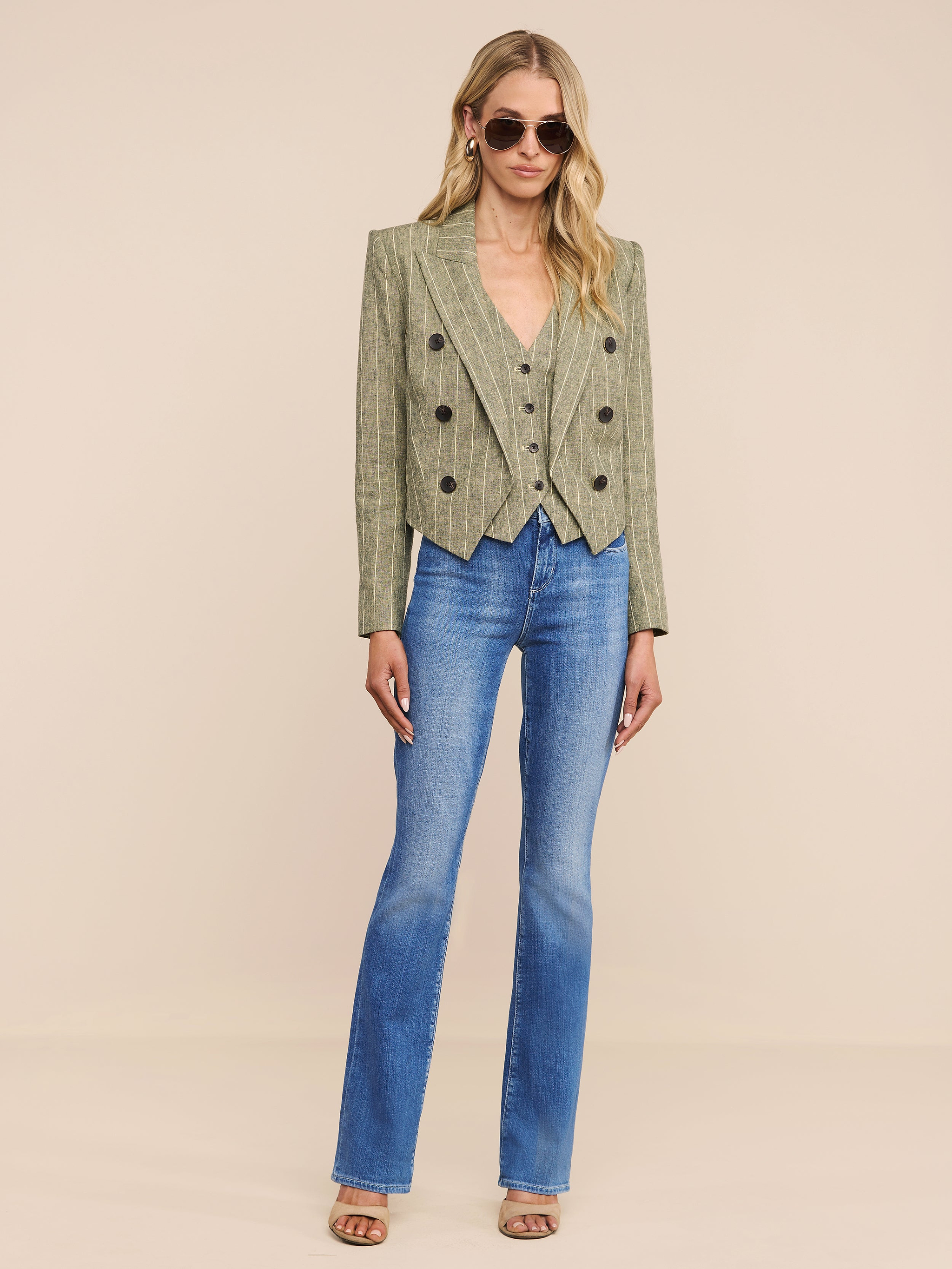 A woman wearing sunglasses, the Lila Linen-Blend Blazer in a cropped green plaid style, blue flared jeans, and open-toe heels stands against a plain white background.