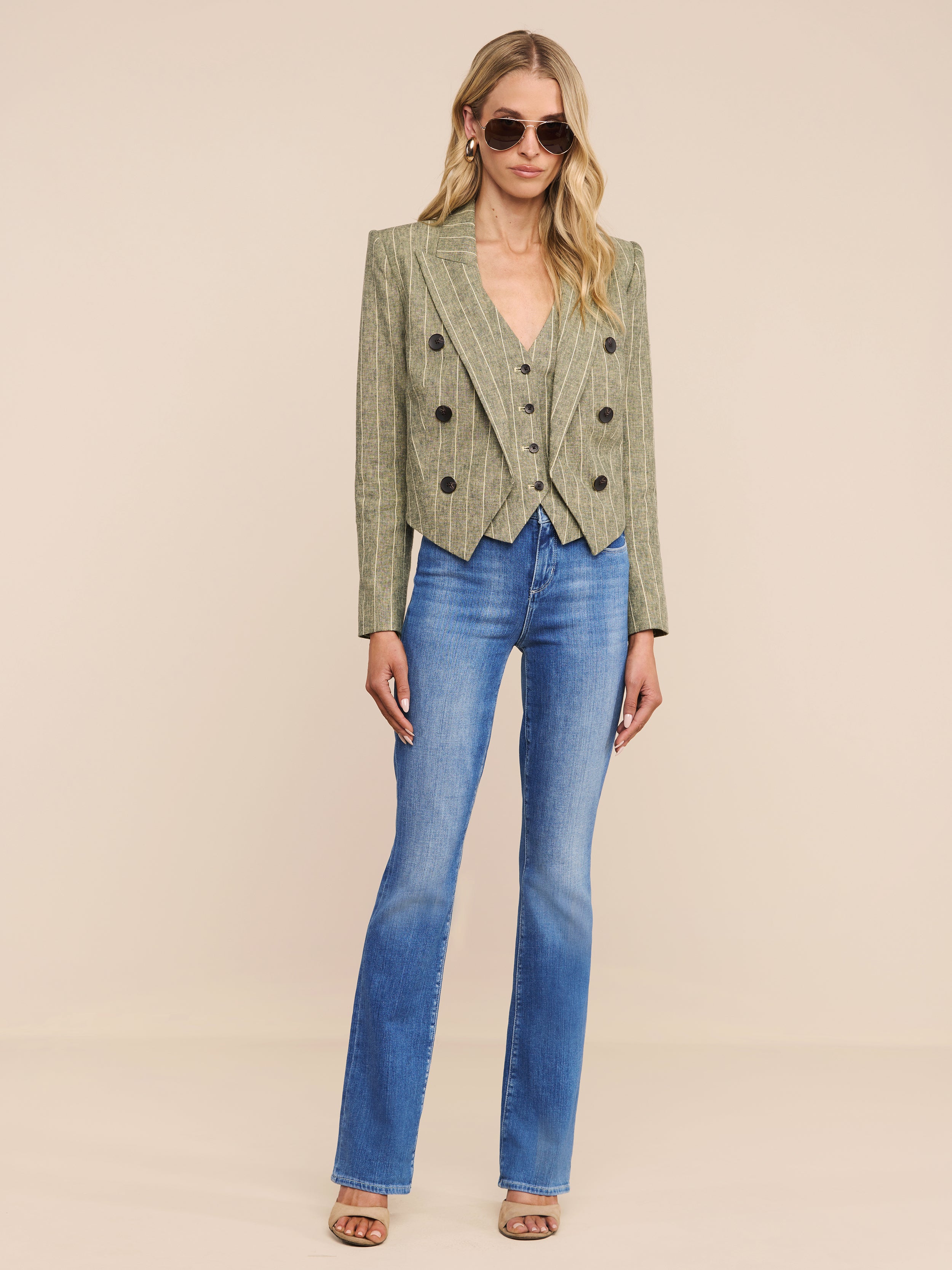 A woman wearing sunglasses, the Lila Linen-Blend Blazer in a cropped green plaid style, blue flared jeans, and open-toe heels stands against a plain white background.