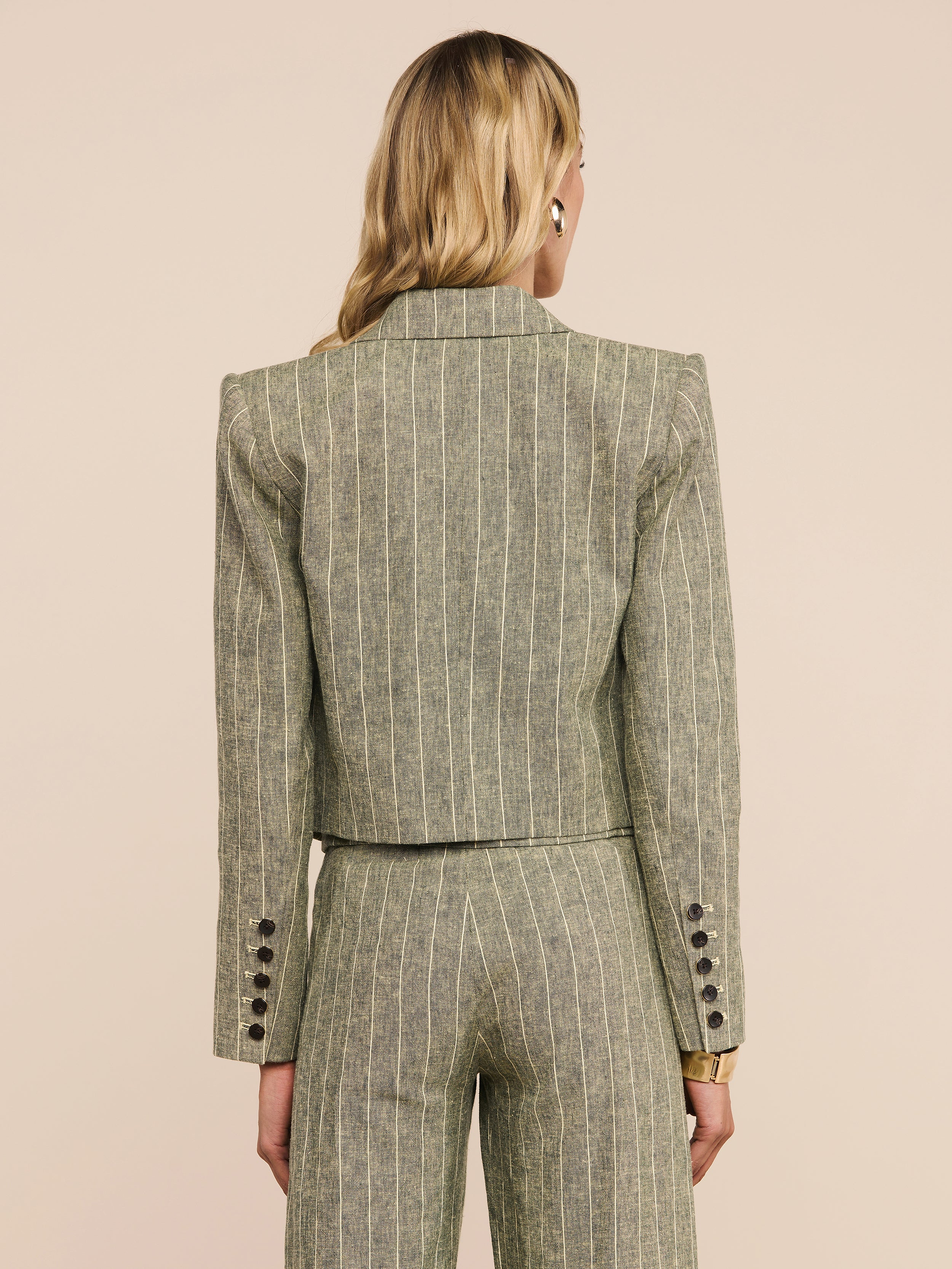 A person with long blonde hair is shown from behind, wearing the Lila Linen-Blend Blazer in a pinstriped olive green design with matching trousers. The suit features button details on the cuffs and offers a refined, fitted silhouette.