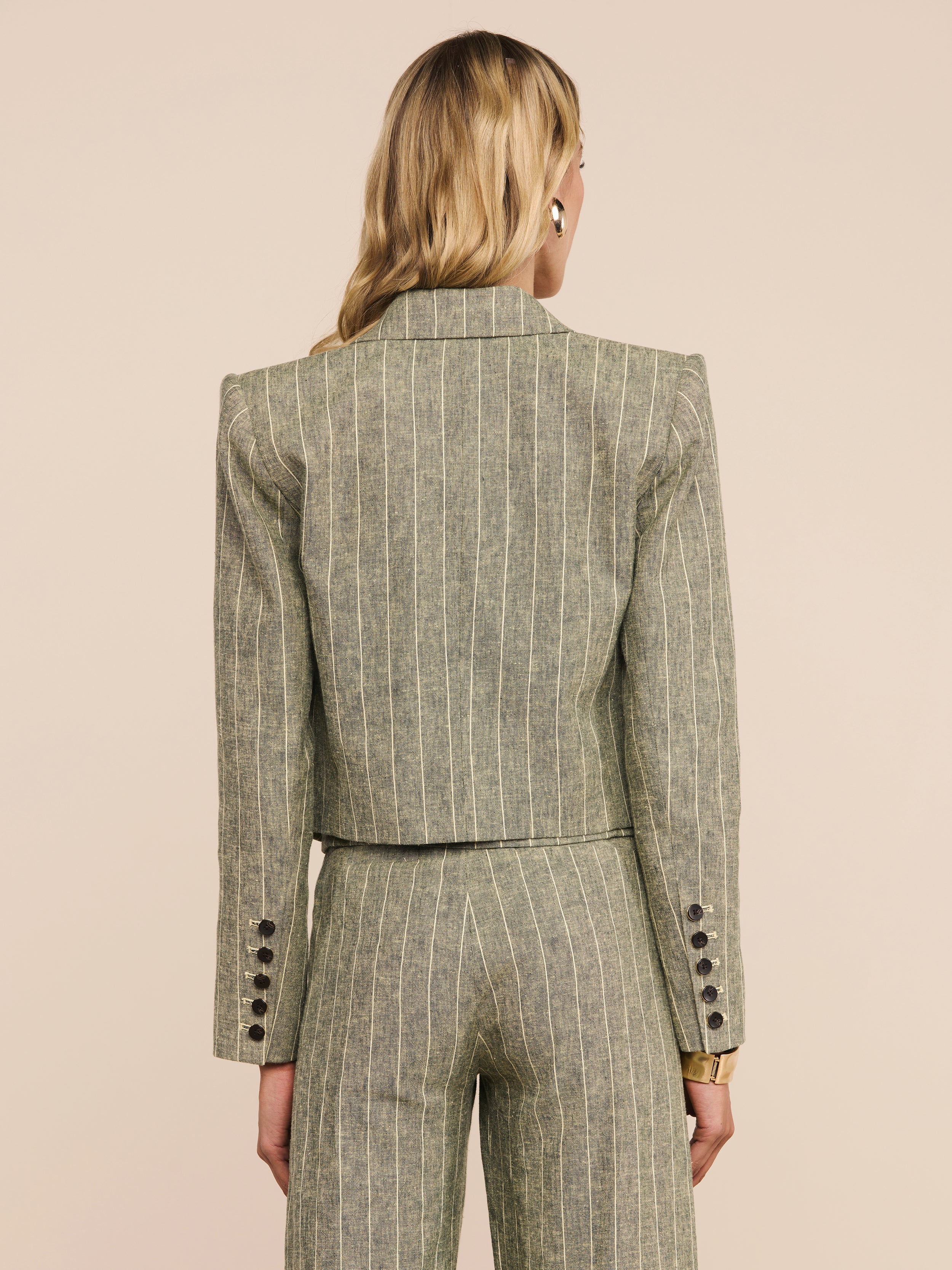 A person with long blonde hair is shown from behind, wearing the Lila Linen-Blend Blazer in a pinstriped olive green design with matching trousers. The suit features button details on the cuffs and offers a refined, fitted silhouette.