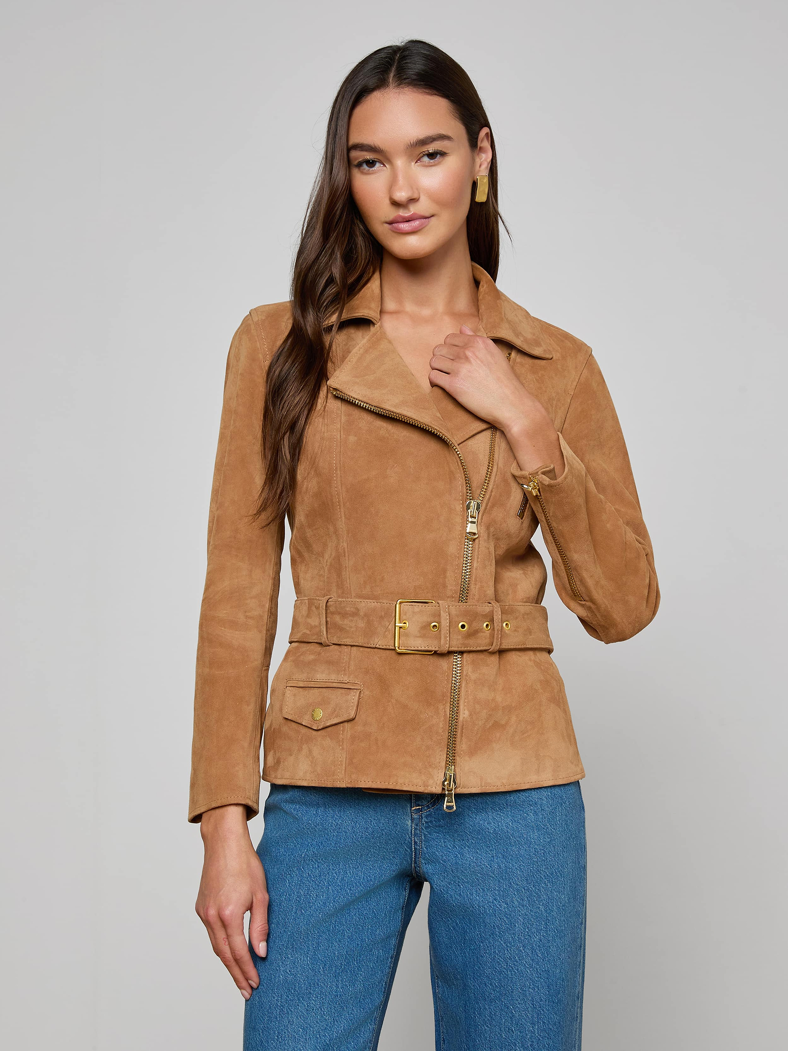 A woman wearing the Dinah Suede Biker Jacket in tan and blue jeans stands against a plain light background, looking at the camera with one hand touching the jacket’s collar. This stylish piece is a standout in women’s outerwear.