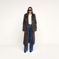 A woman wearing the Lizbeth Faux Fur Jacket—a long, gray shag faux fur coat with a relaxed fit—paired with blue jeans, black shoes, and sunglasses stands against a plain white background.