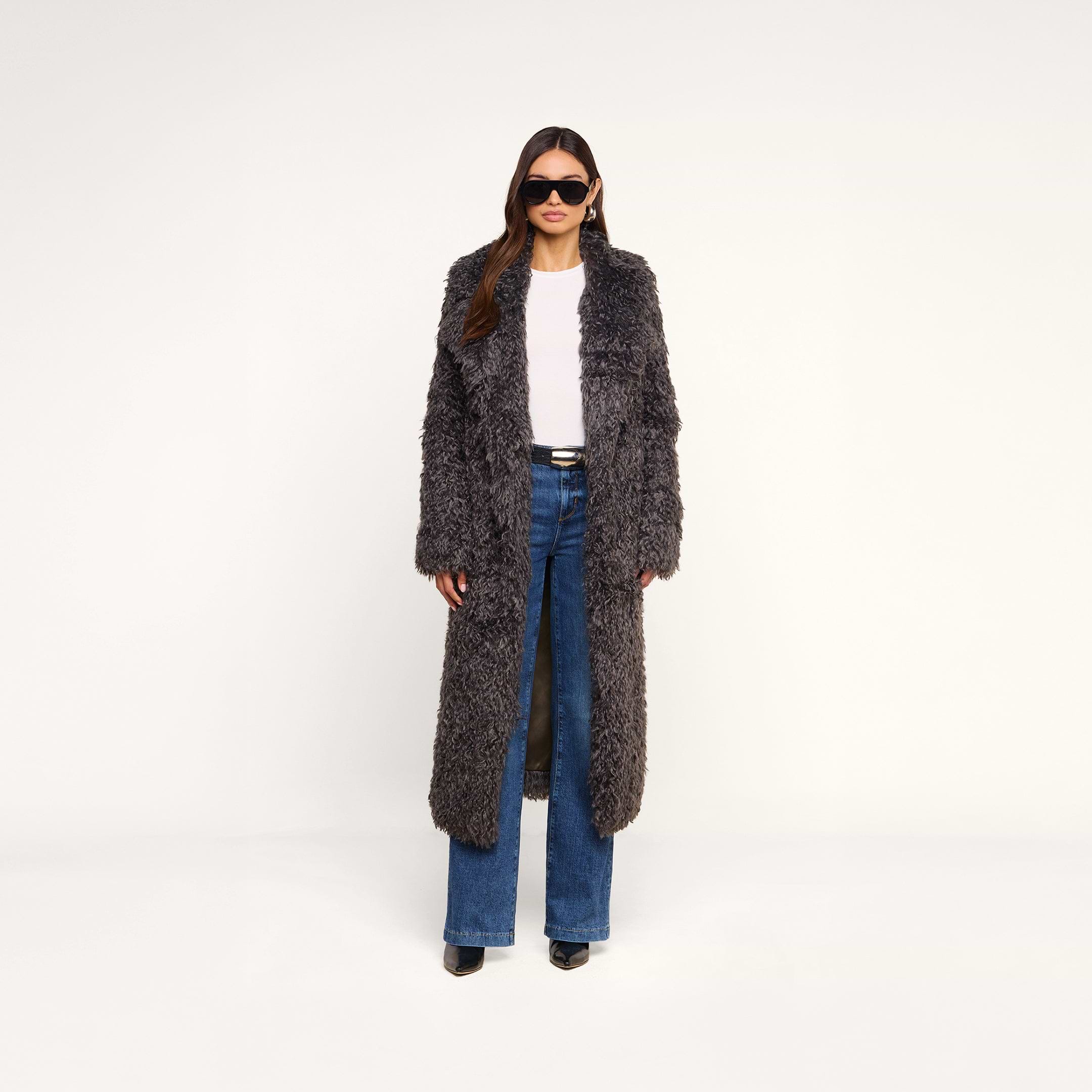 A woman wearing the Lizbeth Faux Fur Jacket—a long, gray shag faux fur coat with a relaxed fit—paired with blue jeans, black shoes, and sunglasses stands against a plain white background.