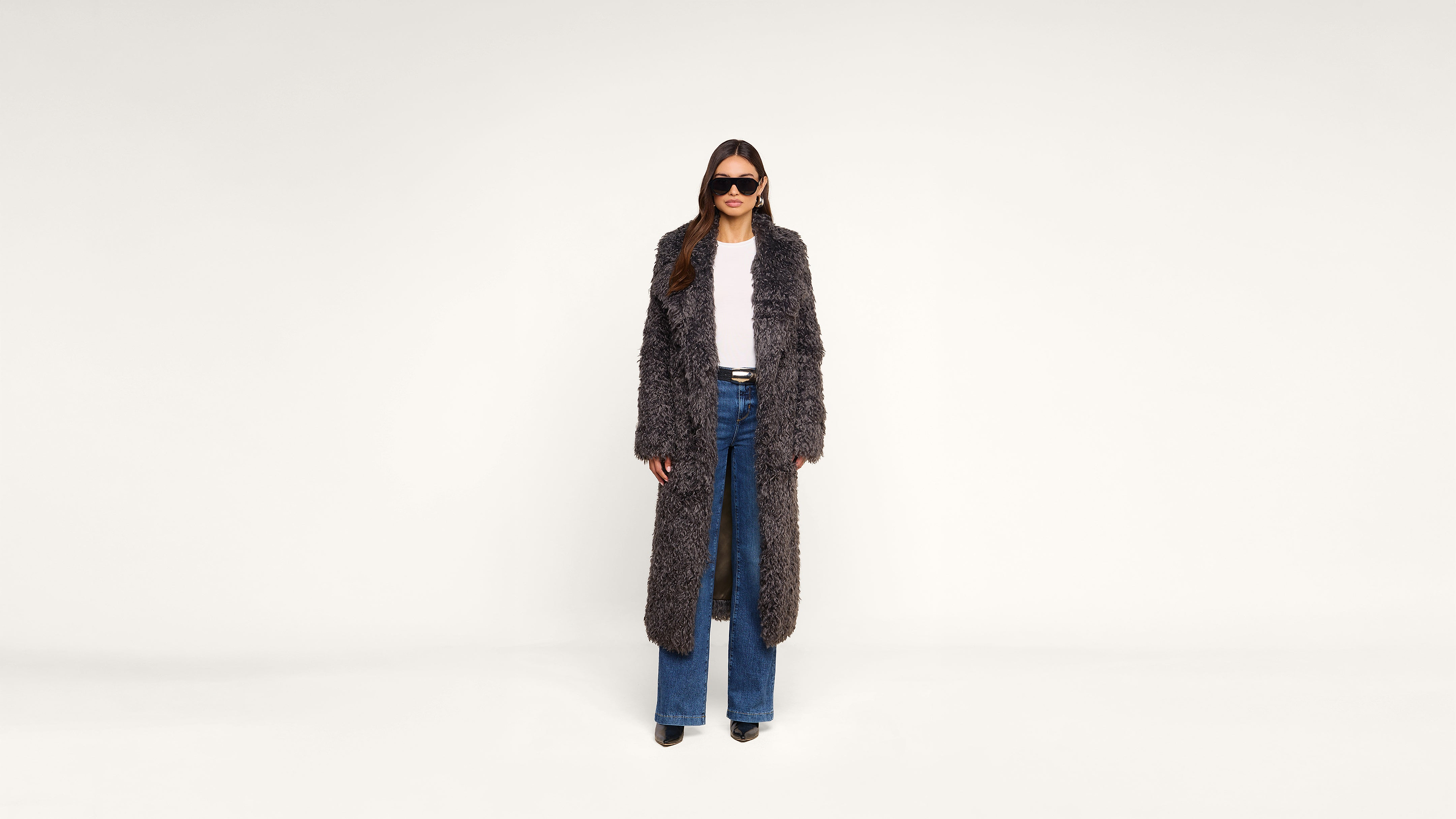 A person wearing sunglasses, a long textured gray coat, white shirt, blue flared jeans, and black shoes stands against a plain white background.