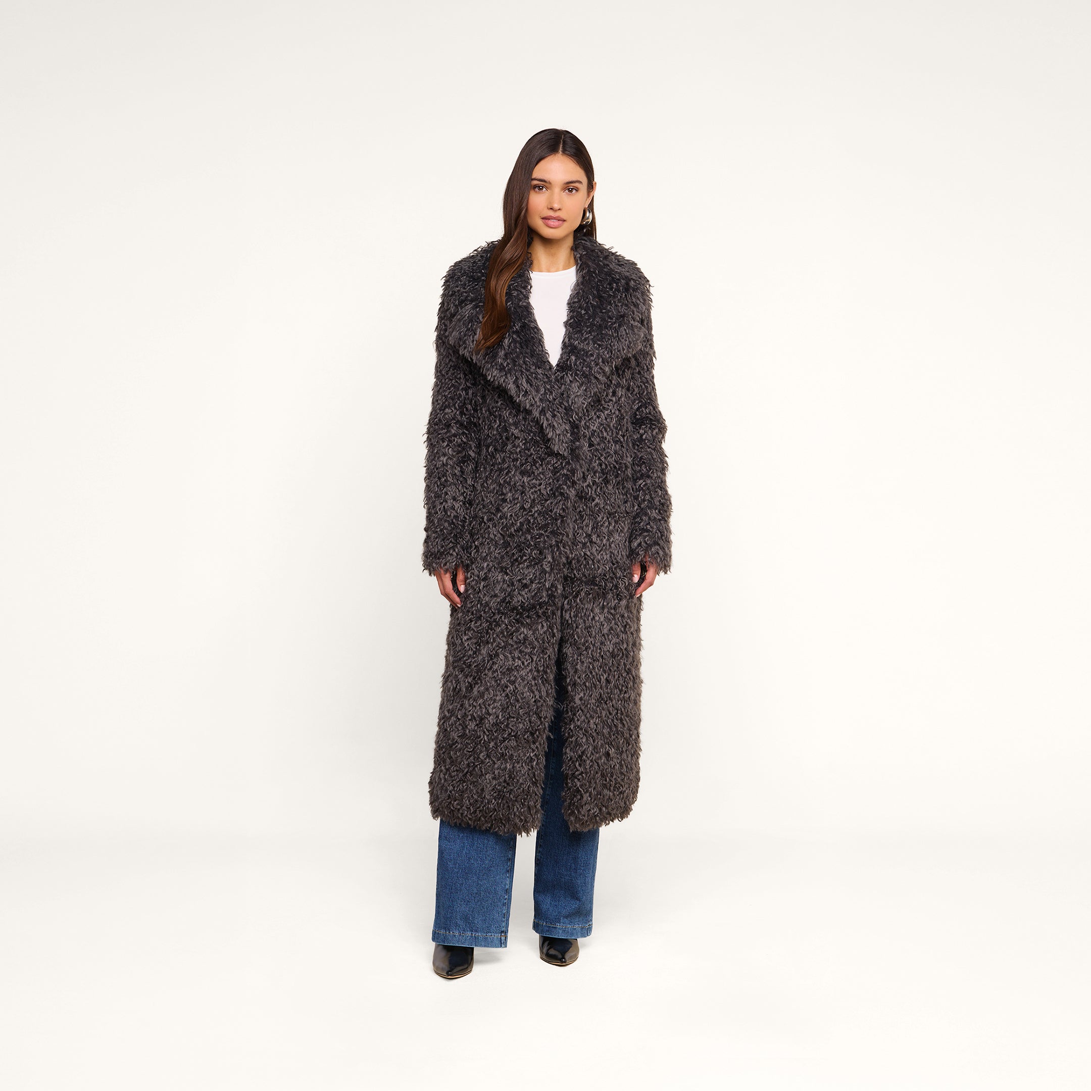 A woman stands facing forward against a plain white background, wearing the Lizbeth Faux Fur Jacket—a long, shag faux fur gray coat—layered over a white top and blue jeans, finished with black heeled shoes.