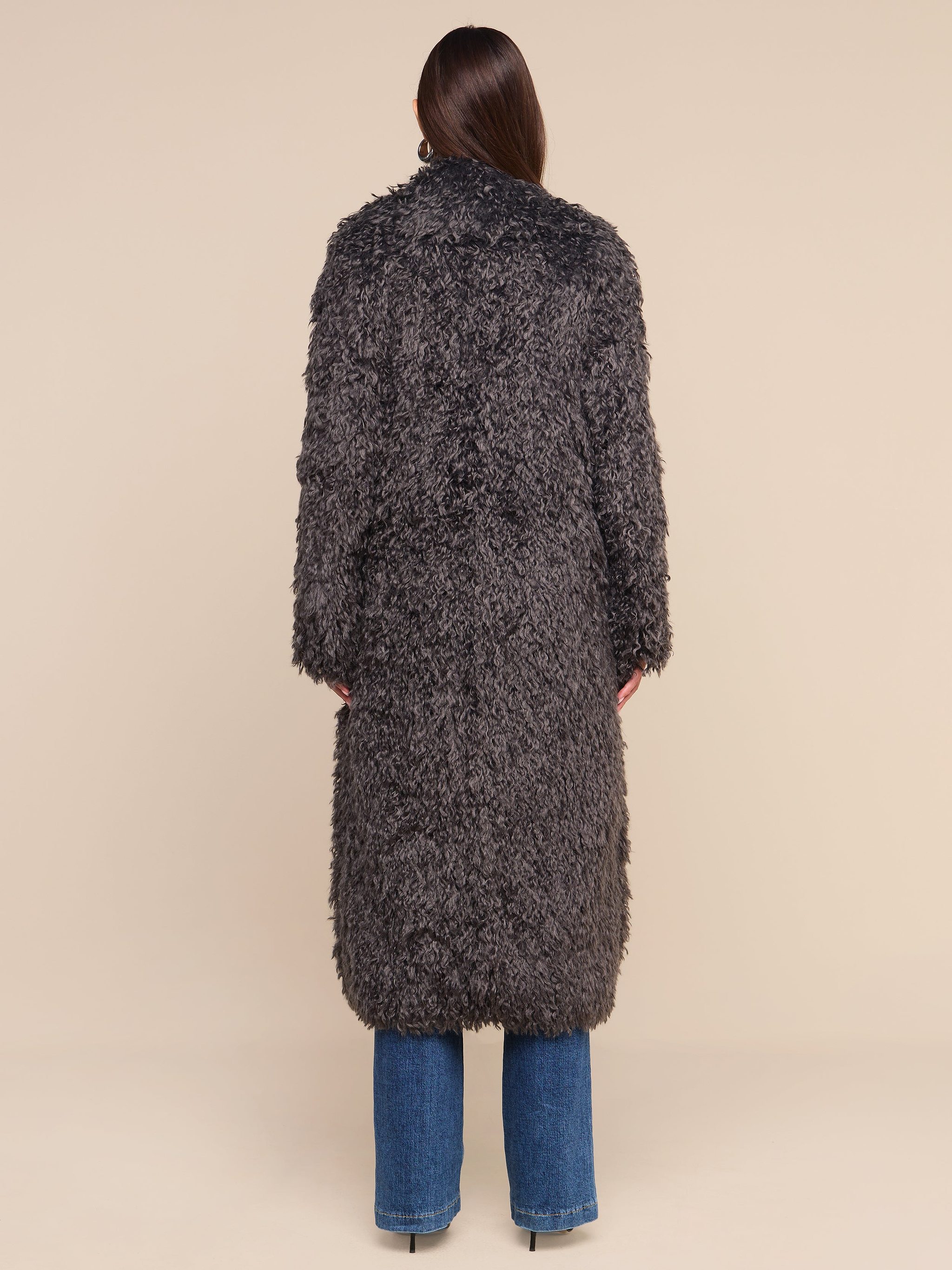 A person stands facing away, wearing the Lizbeth Faux Fur Jacket—a long, textured dark gray shag coat—paired with blue jeans against a plain white background.