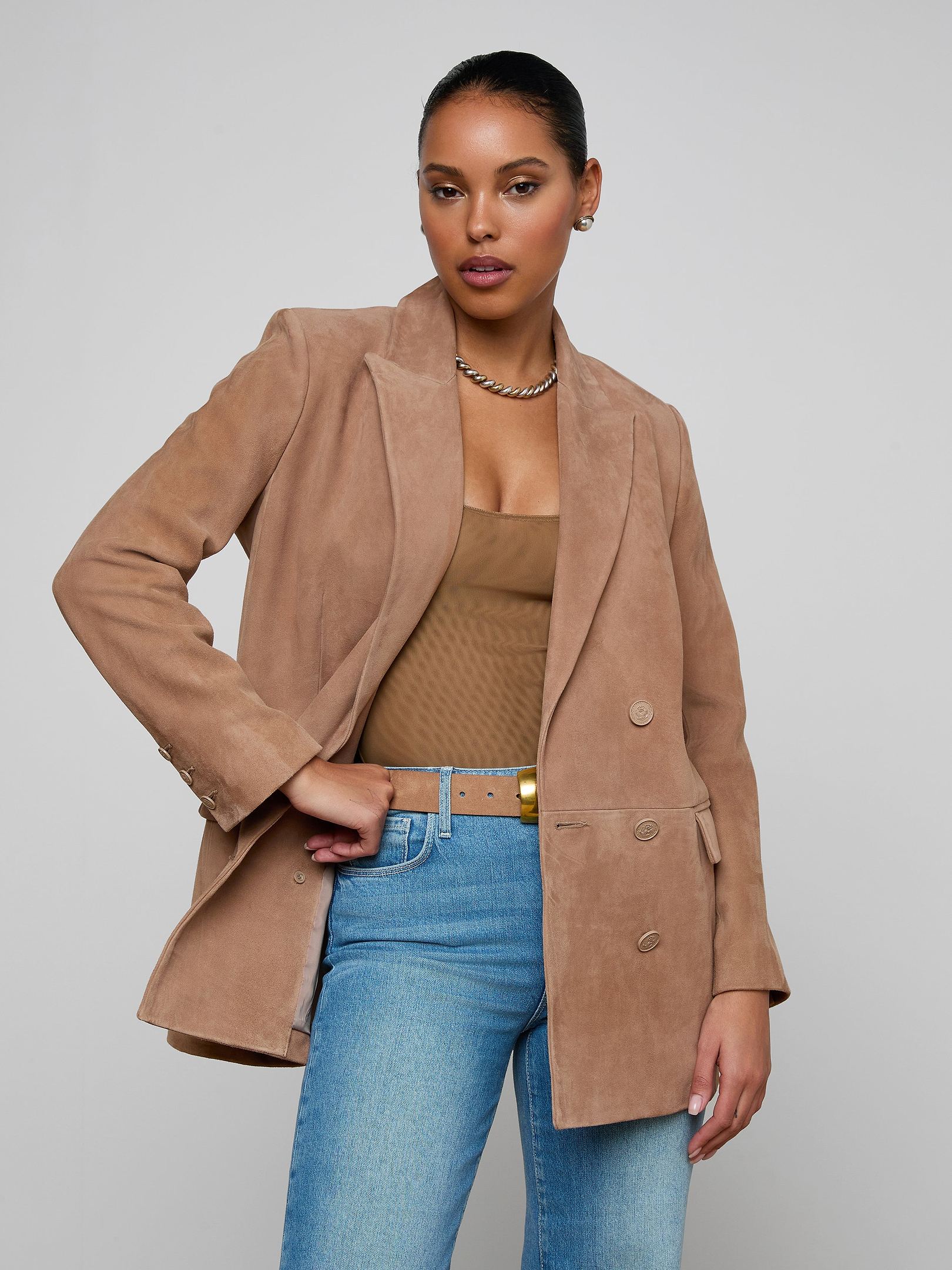 Wearing the Aimee Oversized Suede Blazer in tan over a brown top and blue jeans, accessorized with a gold necklace, a woman stands against a plain light background, gazing confidently at the camera.